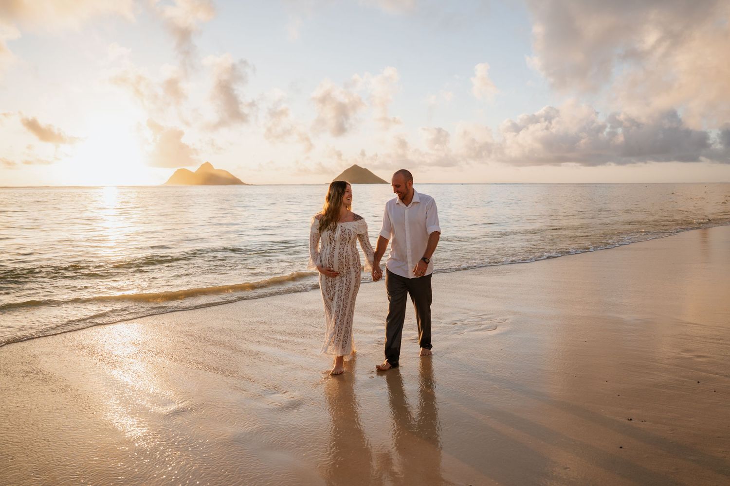 Couple holding hands on a golden sandy beach at sunset with ocean waves and island silhouette in background.