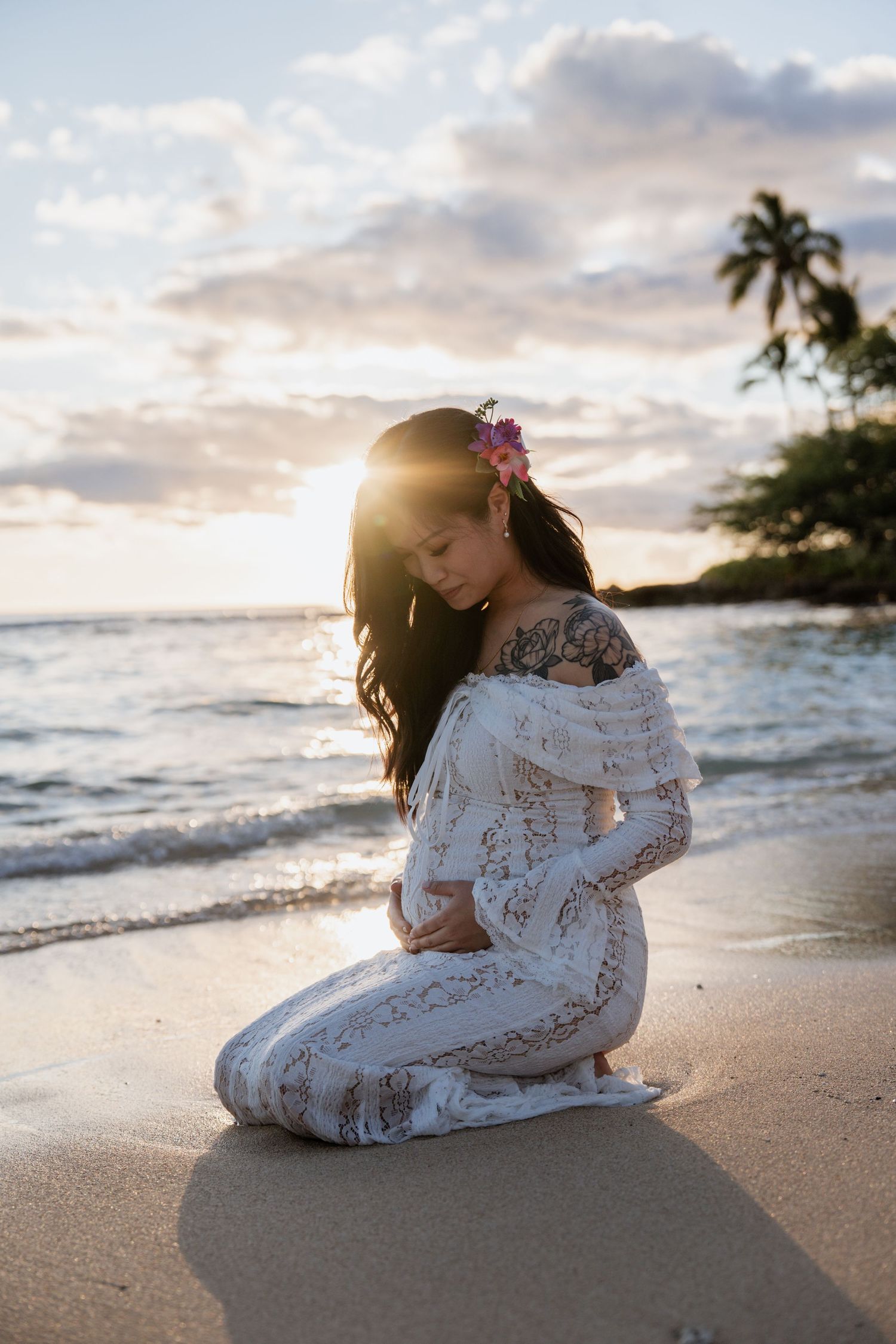 A tender moment captured between mother and child sitting on a tropical beach at sunset in white lace dresses.