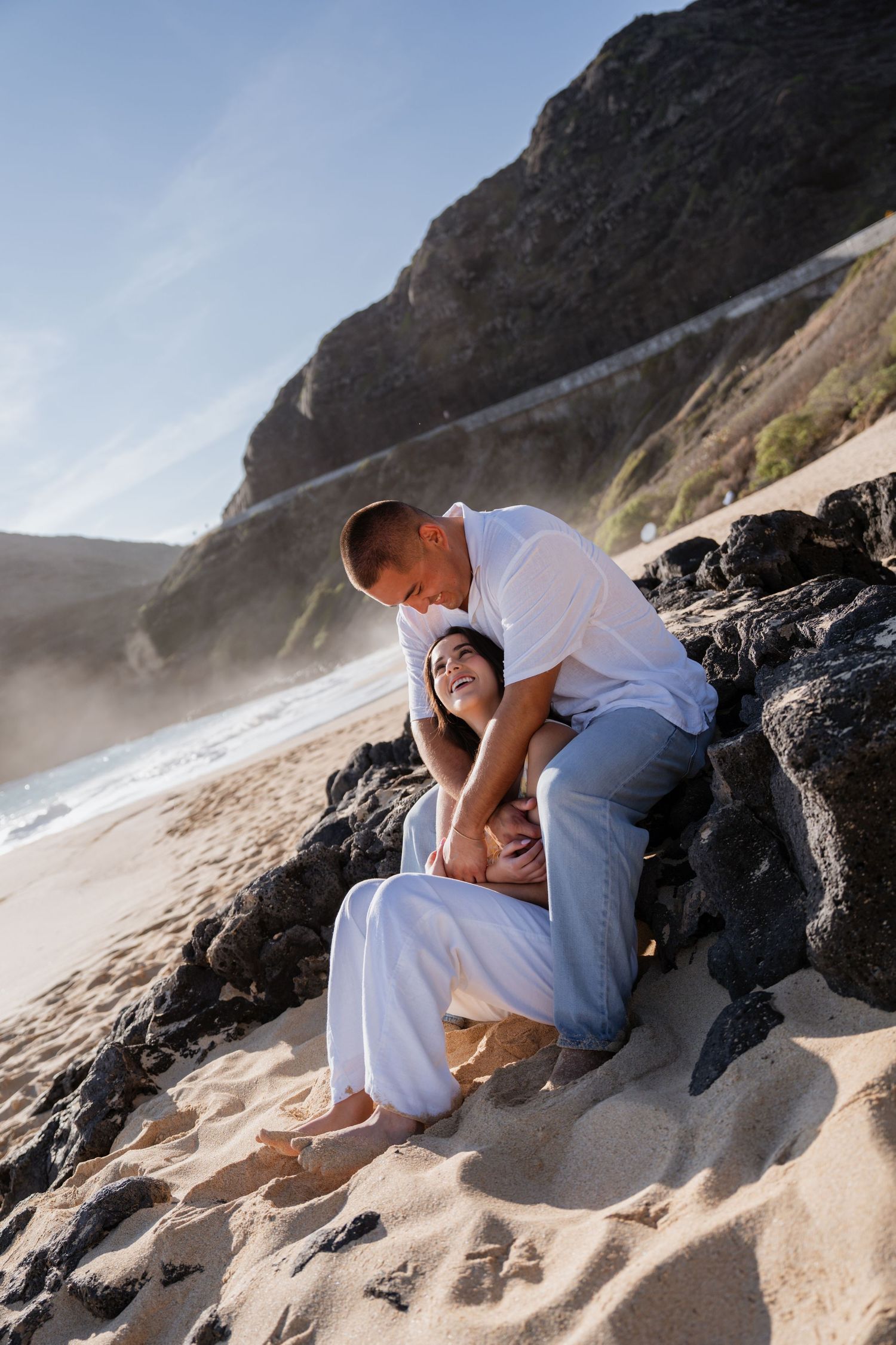 Romantic couple embracing on sandy beach with dramatic cliffs and ocean waves in background during sunset photoshoot. Waikiki photographer