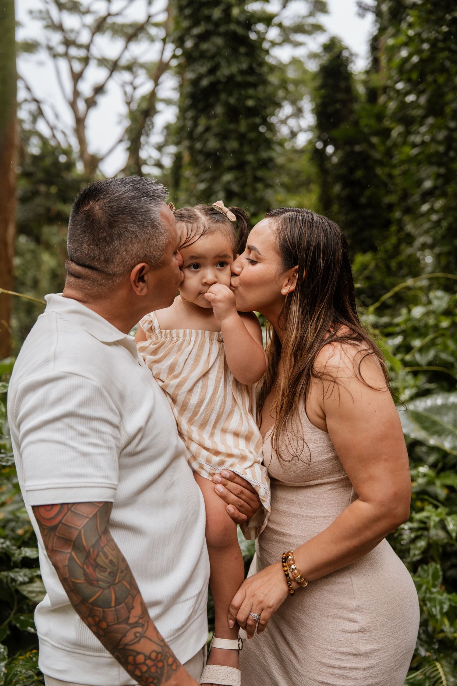 Parents kissing their toddler daughter outdoors in a lush green garden setting.
