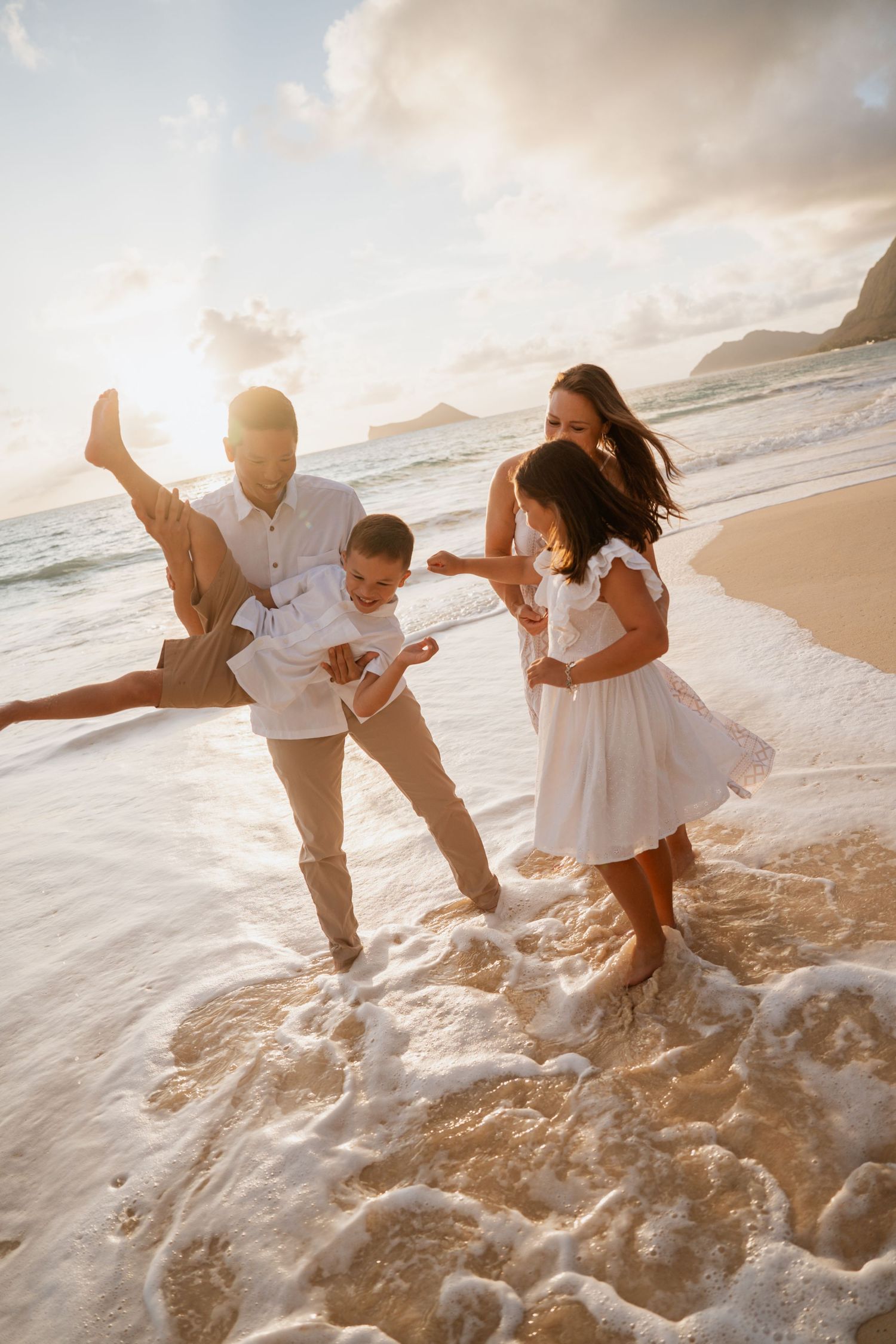 Family plays together in ocean waves at sunset on a tropical beach during golden hour photo session.