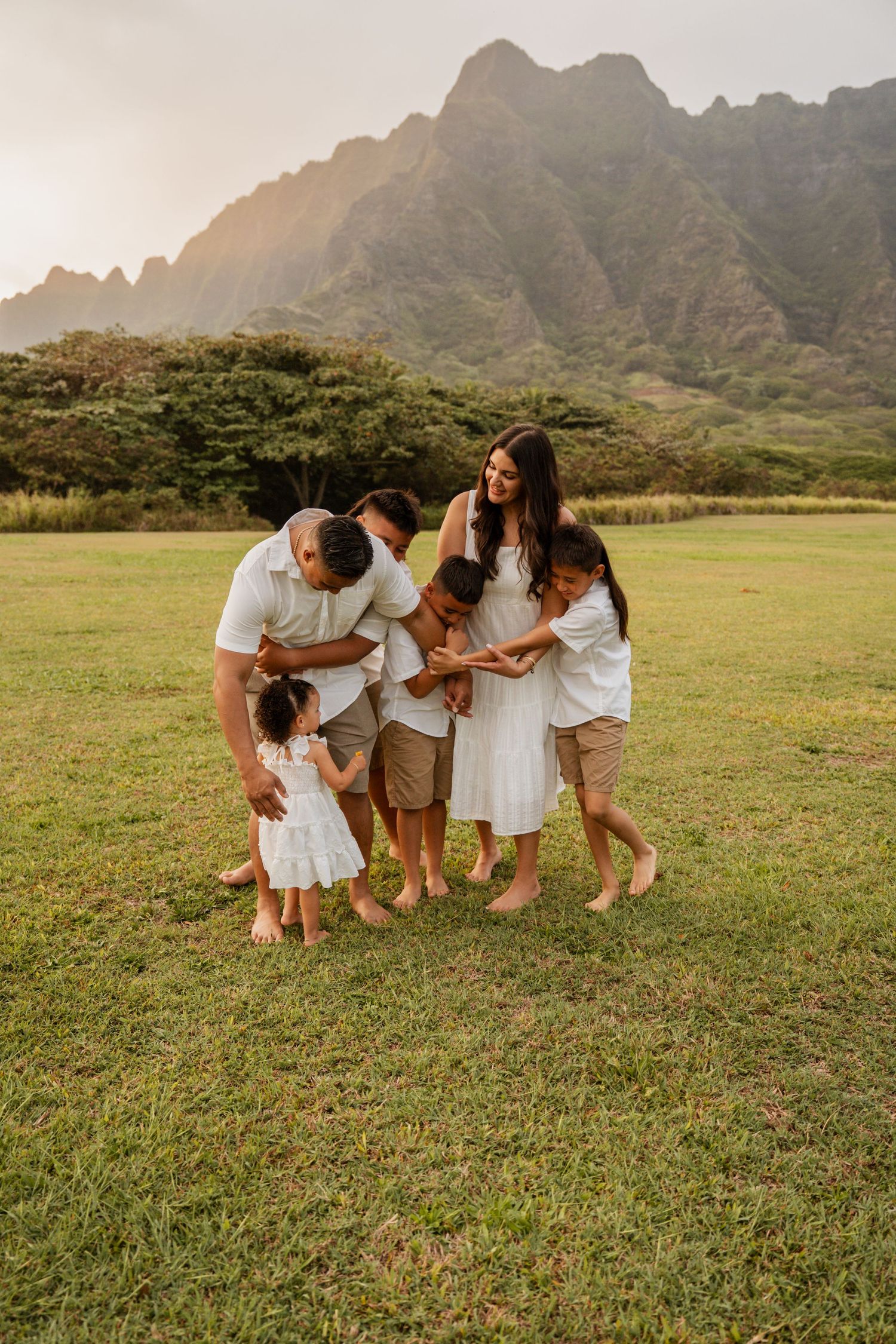 Family of six sharing a joyful embrace on a green lawn with dramatic Hawaiian mountains at sunset.