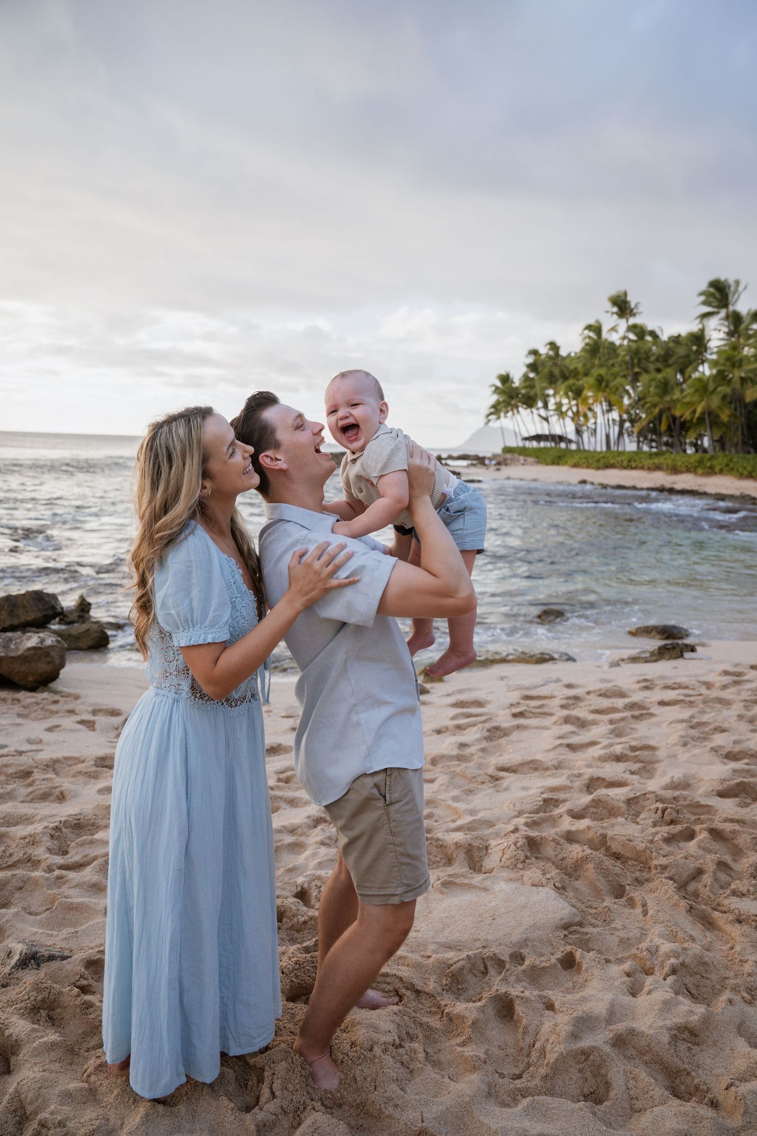 A family shares tender moments together on a rocky beach with palm trees and ocean waves in the background. Disney resort Hawaii, Aulani