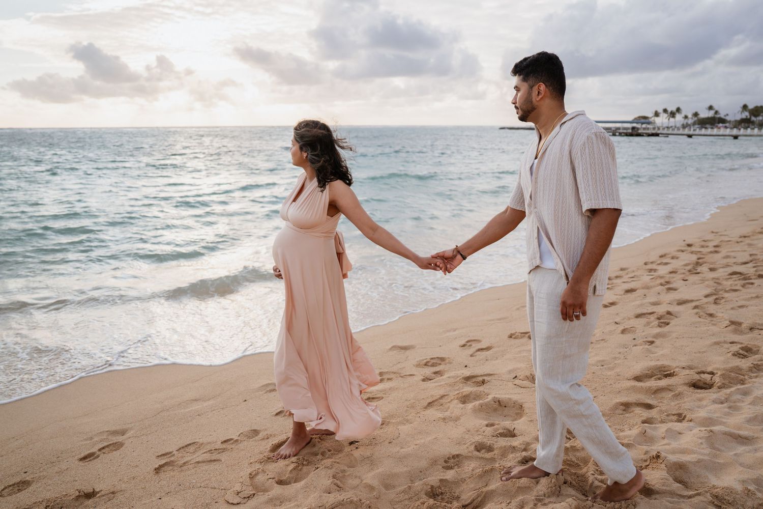 Pregnant couple holding hands walking on sandy beach at sunset, woman in pink dress, man in white outfit.