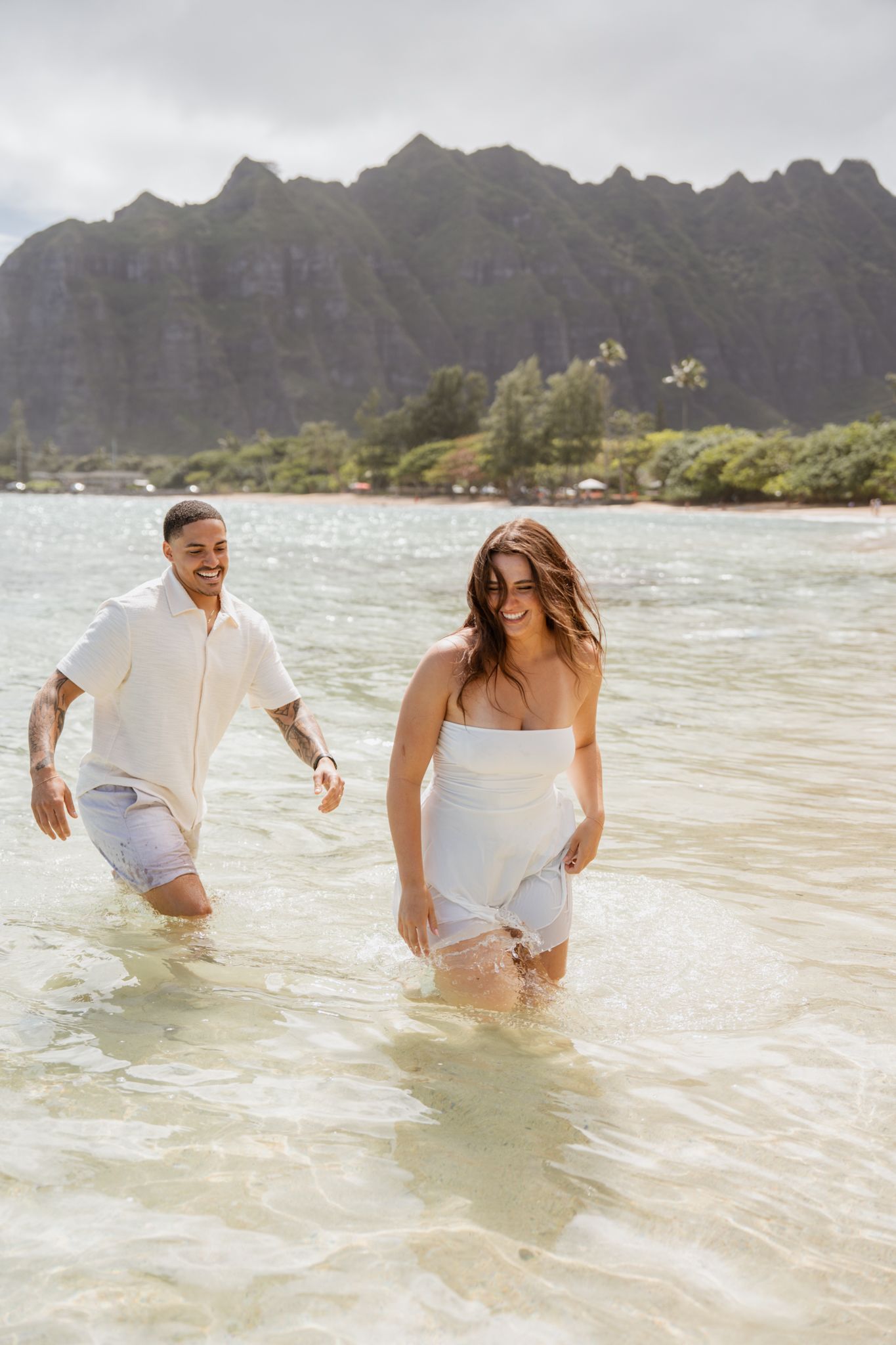 A couple wades through crystal clear waters with dramatic mountain peaks in the background at a Hawaiian beach.