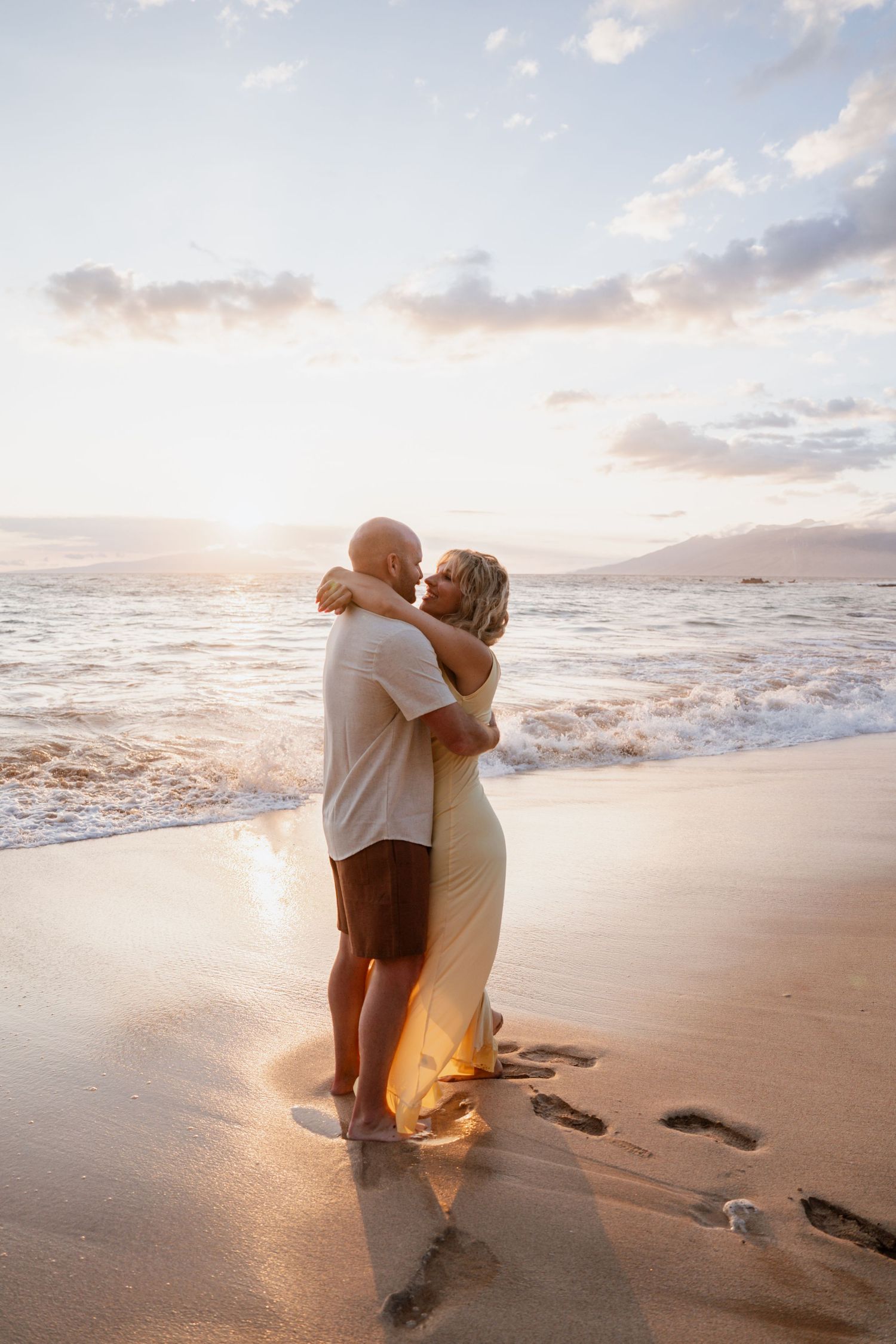 Couple embracing on a sandy beach at sunset, with ocean waves and golden light creating a romantic scene.