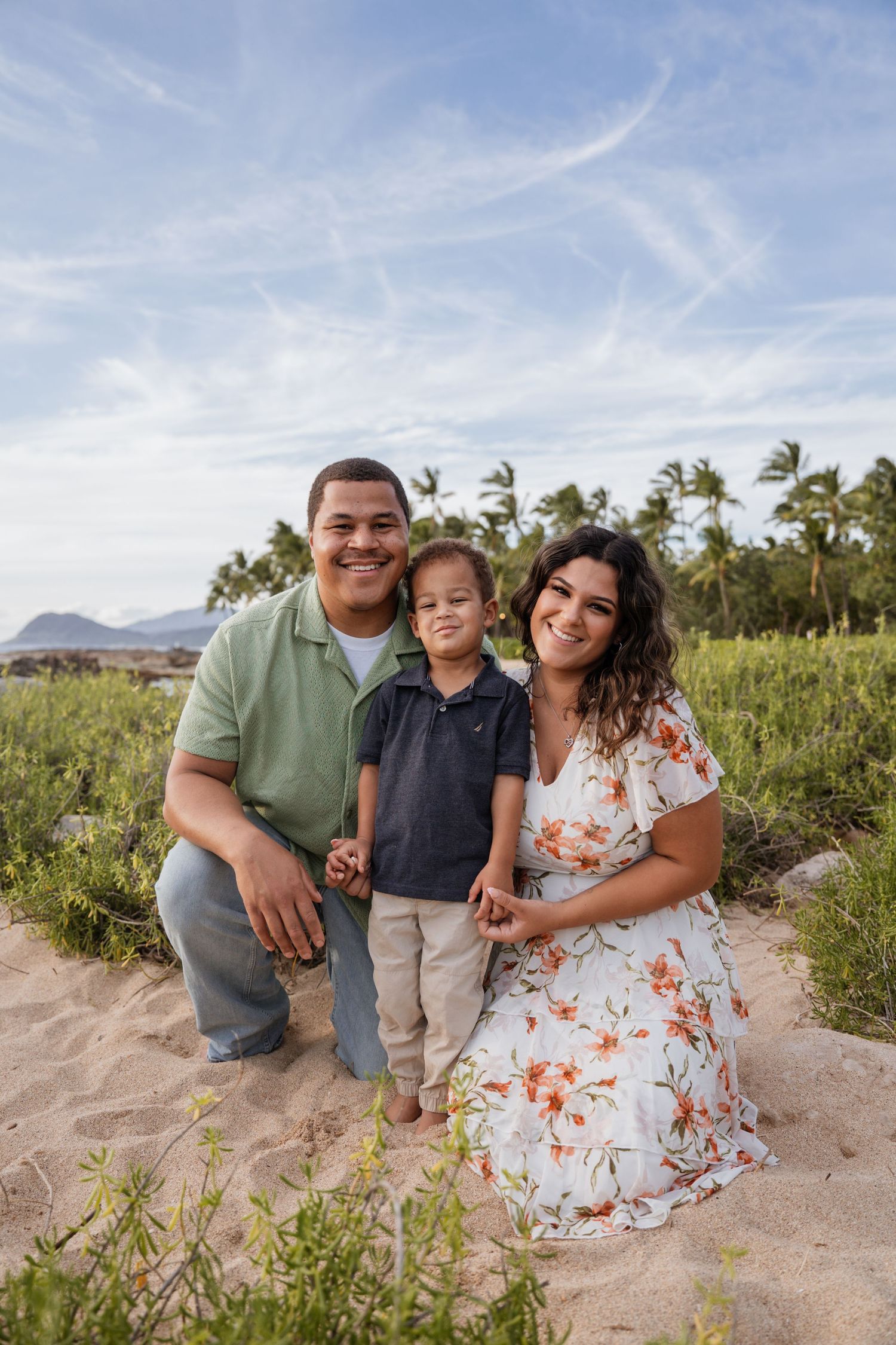Family of three smiling on a sandy beach with palm trees and mountains in Hawaii.