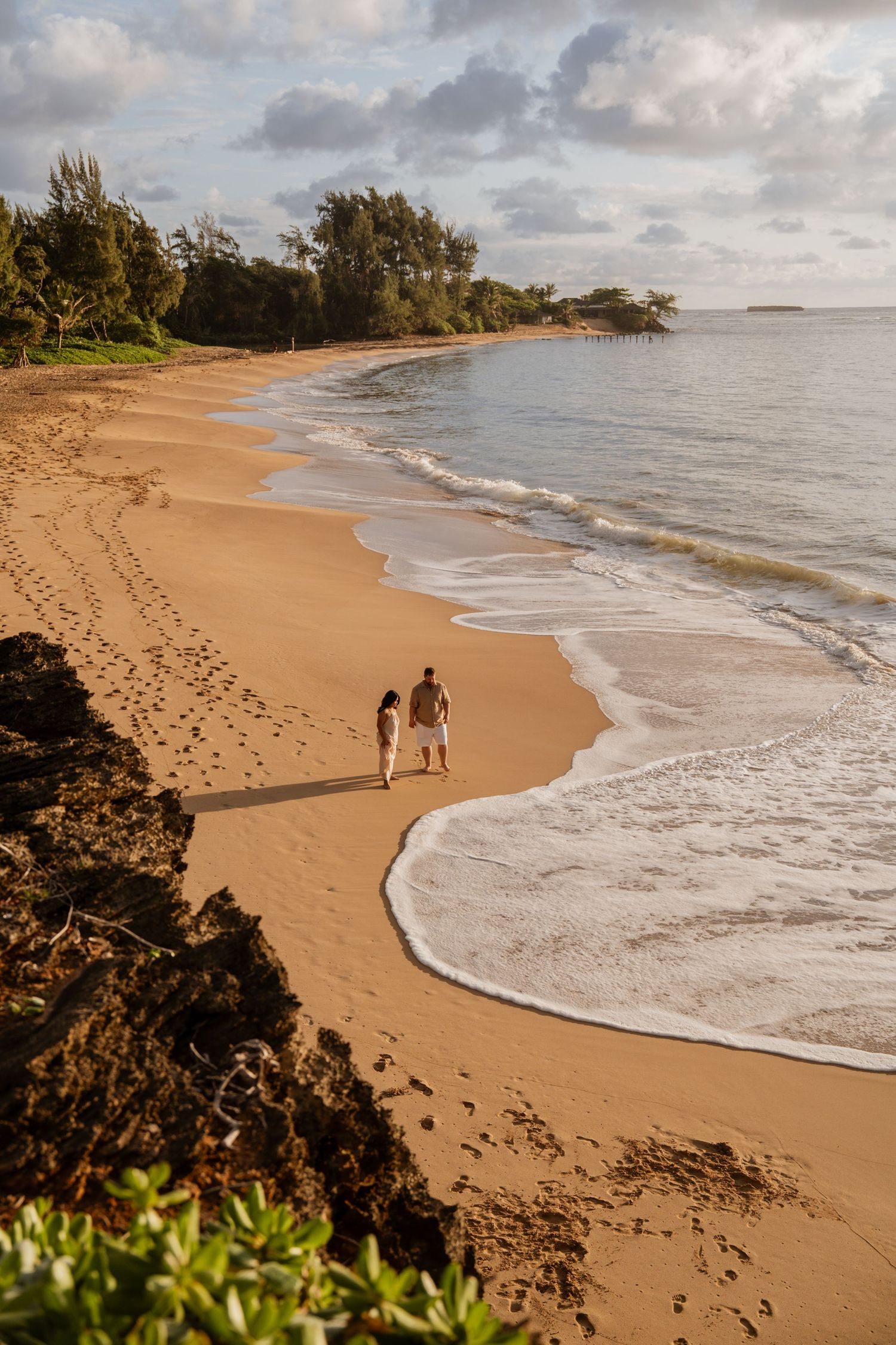 Couple walking hand in hand along a golden sandy beach at sunset with gentle waves and lush green trees in the background.