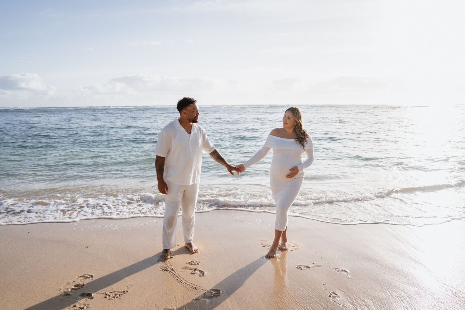 Couple in white outfits holding hands while walking along sunny beach shoreline.