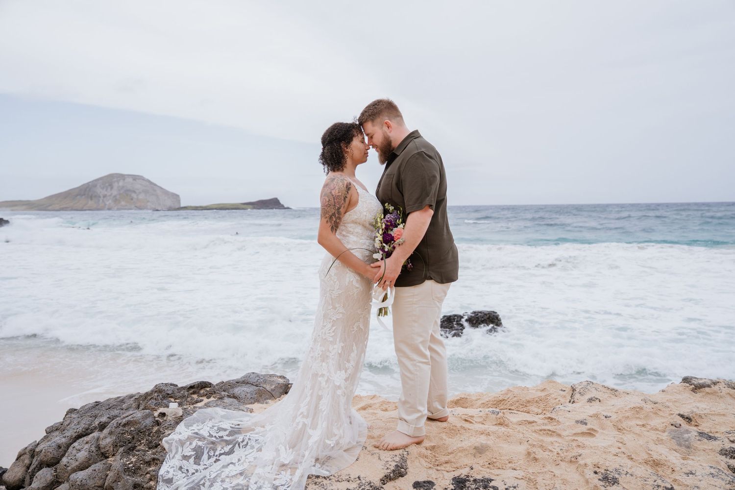 A romantic beach wedding ceremony takes place along the rocky Hawaiian coastline with crashing waves and mountains in the distance.