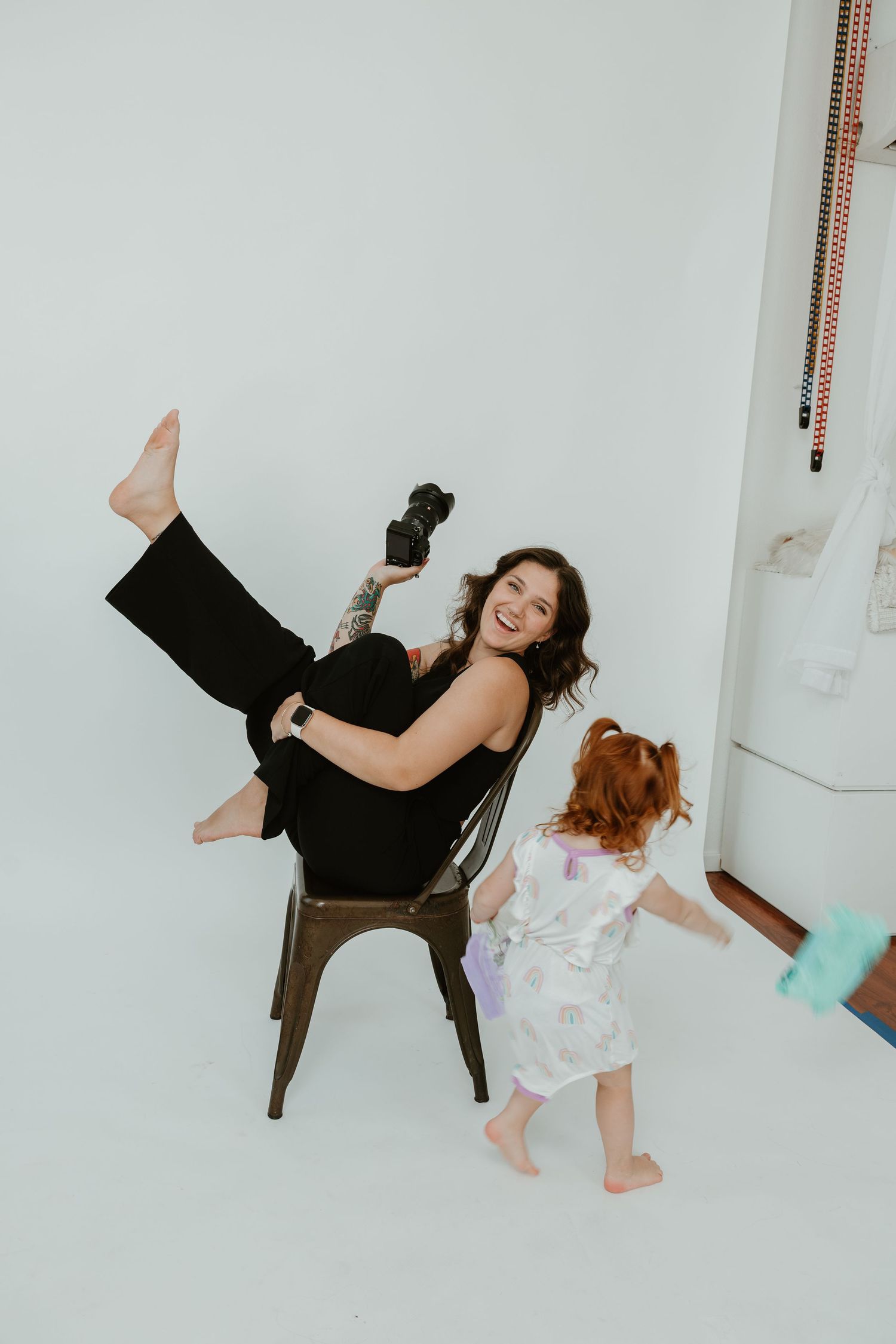 A playful studio photoshoot with a person in black sitting on a chair with leg raised as a child dances nearby.