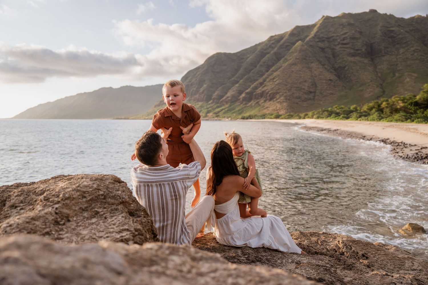 Family sitting on rocks by a scenic Hawaiian beach with lush green mountains in the background.