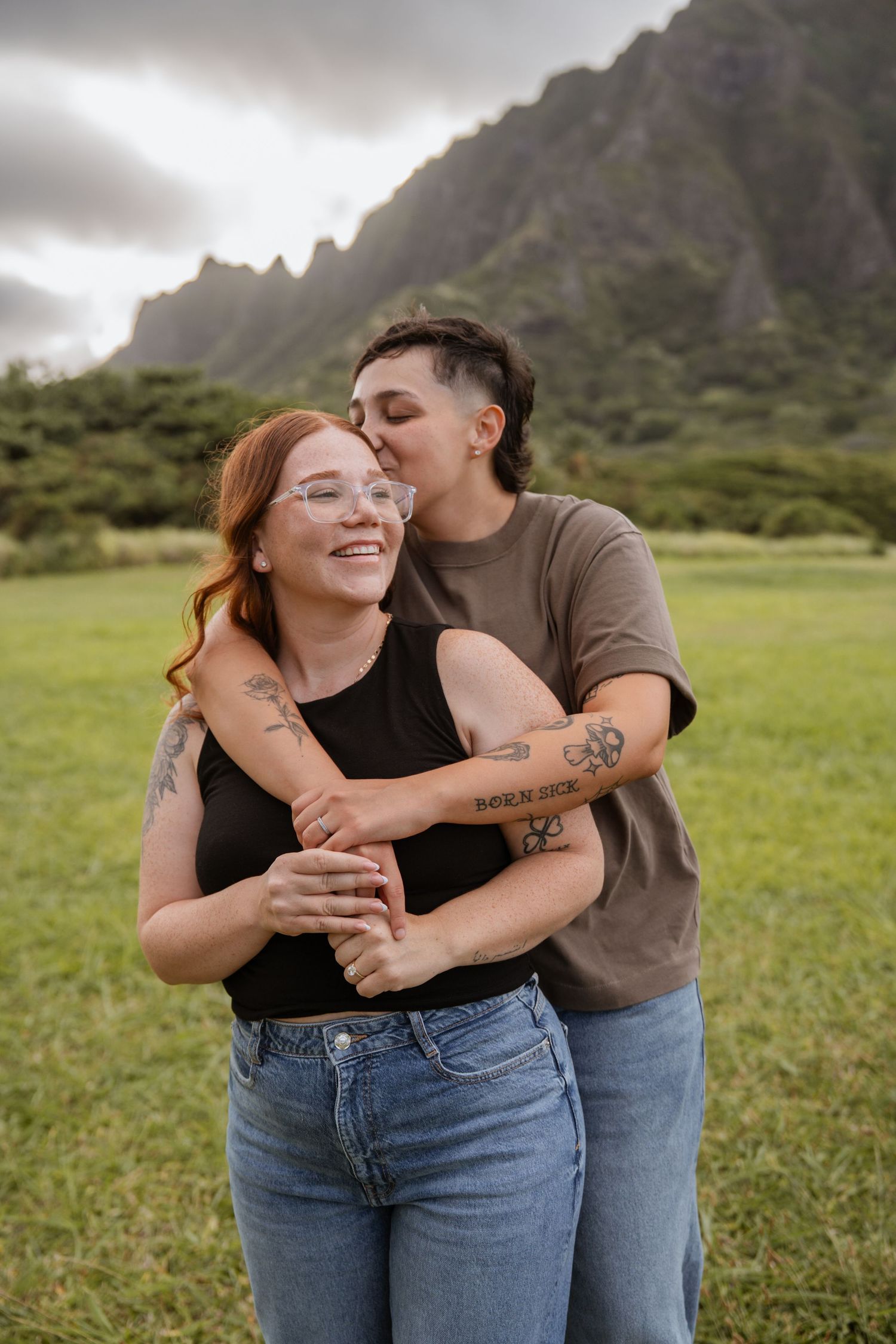 A loving couple embraces in a grassy field with dramatic mountains rising in the background in Hawaii.