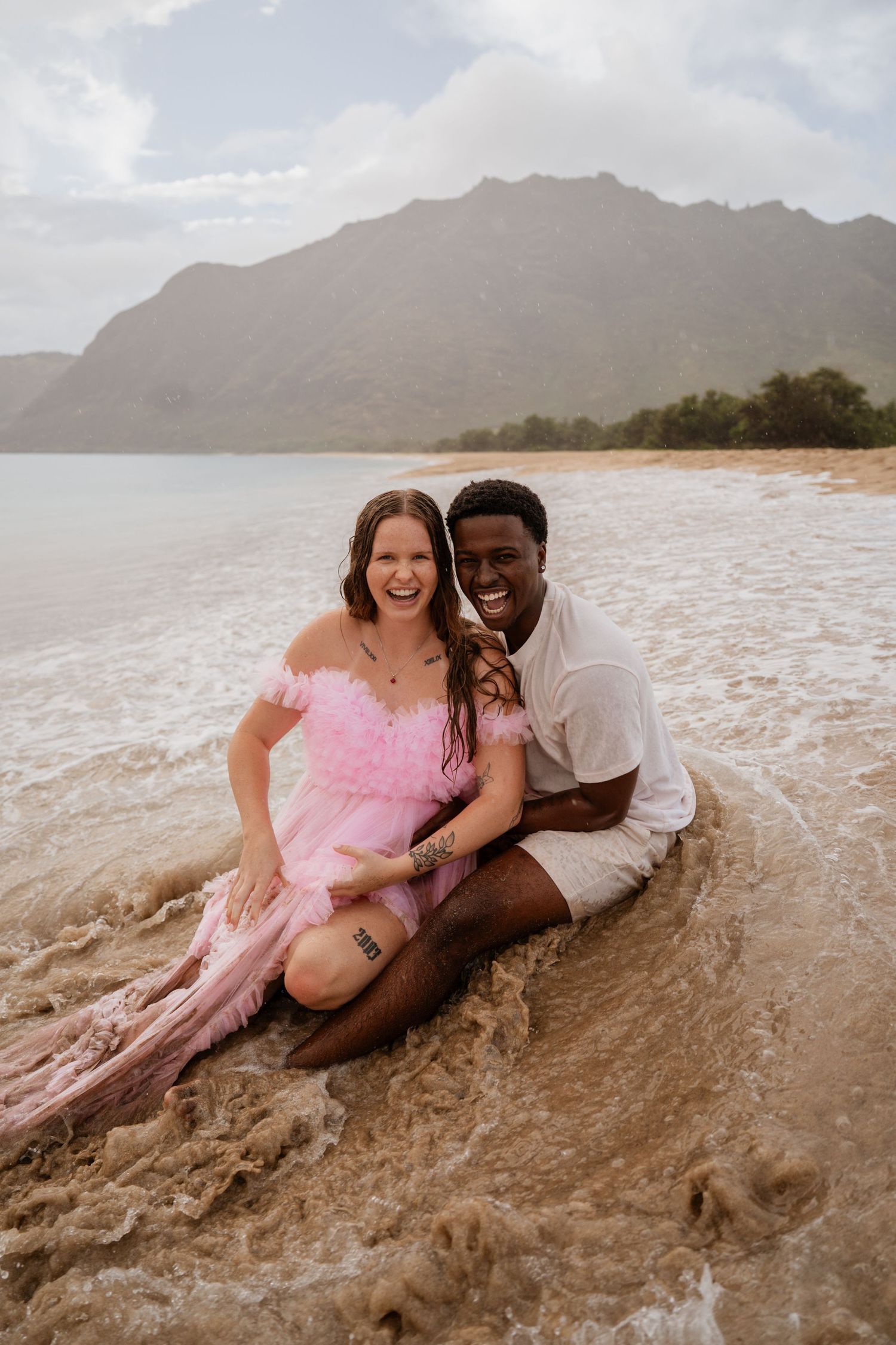 A couple in casual beach attire share a playful moment laying in the sand on a Hawaiian beach at sunset.