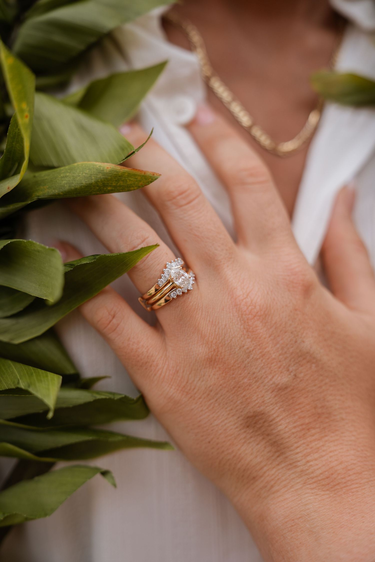 A close-up of a hand wearing stacked gold and diamond rings next to green leaves, with a gold chain necklace visible.