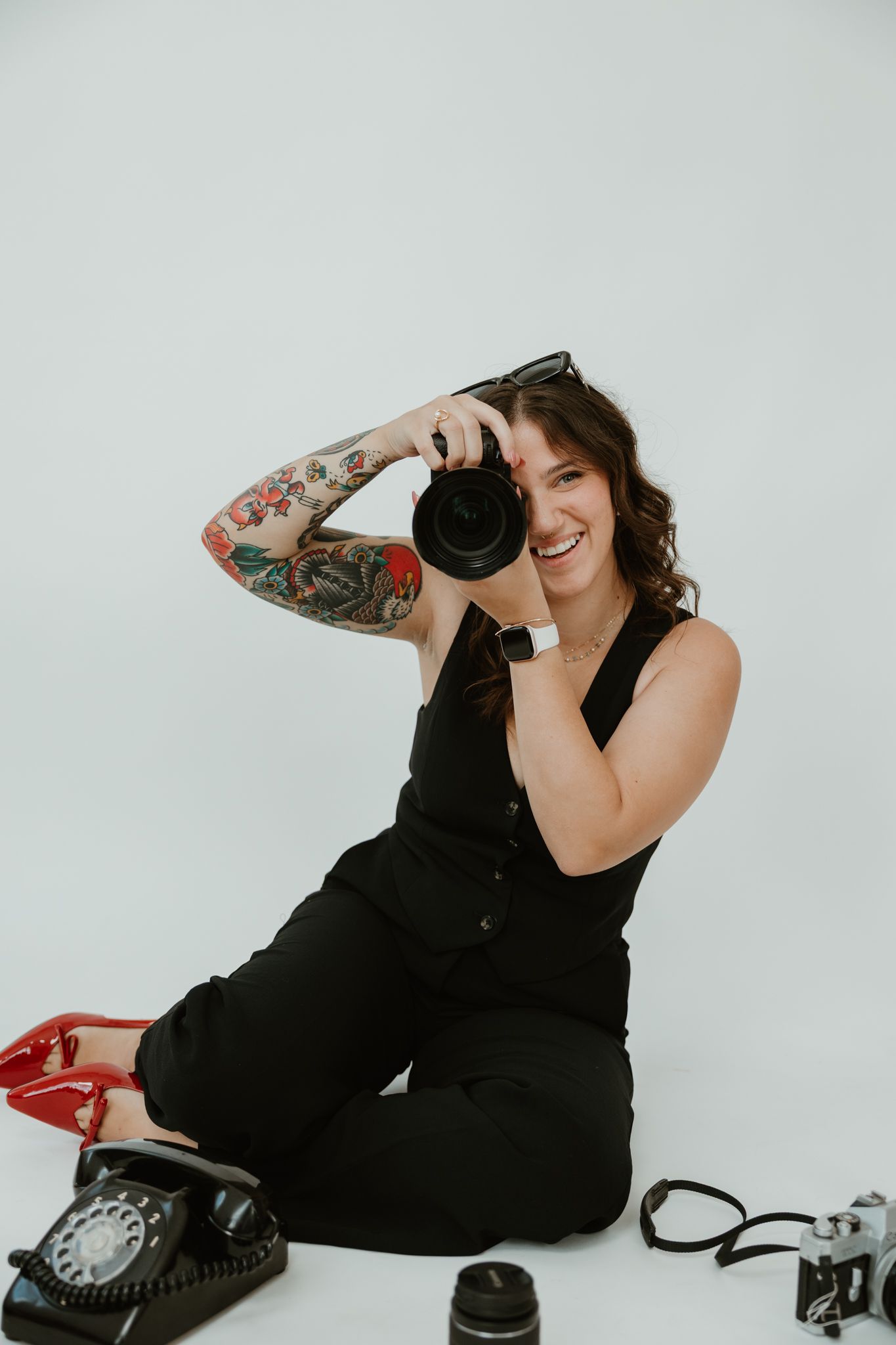 Photographer with colorful tattoos and camera laughs while sitting against white backdrop.