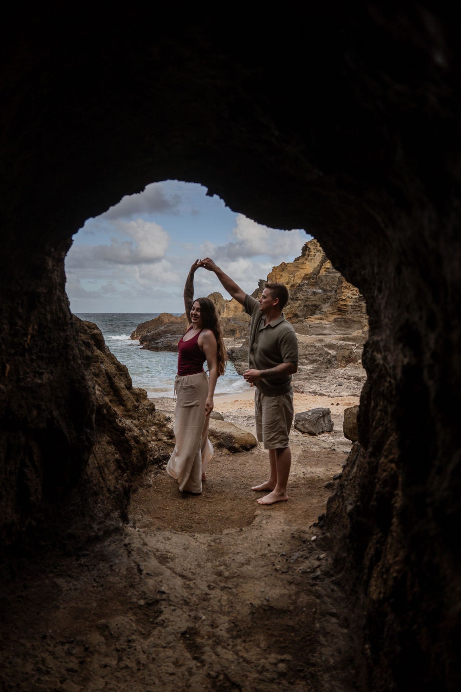 Couple dancing in a cave opening with ocean waves and rocky coastline visible in the background.