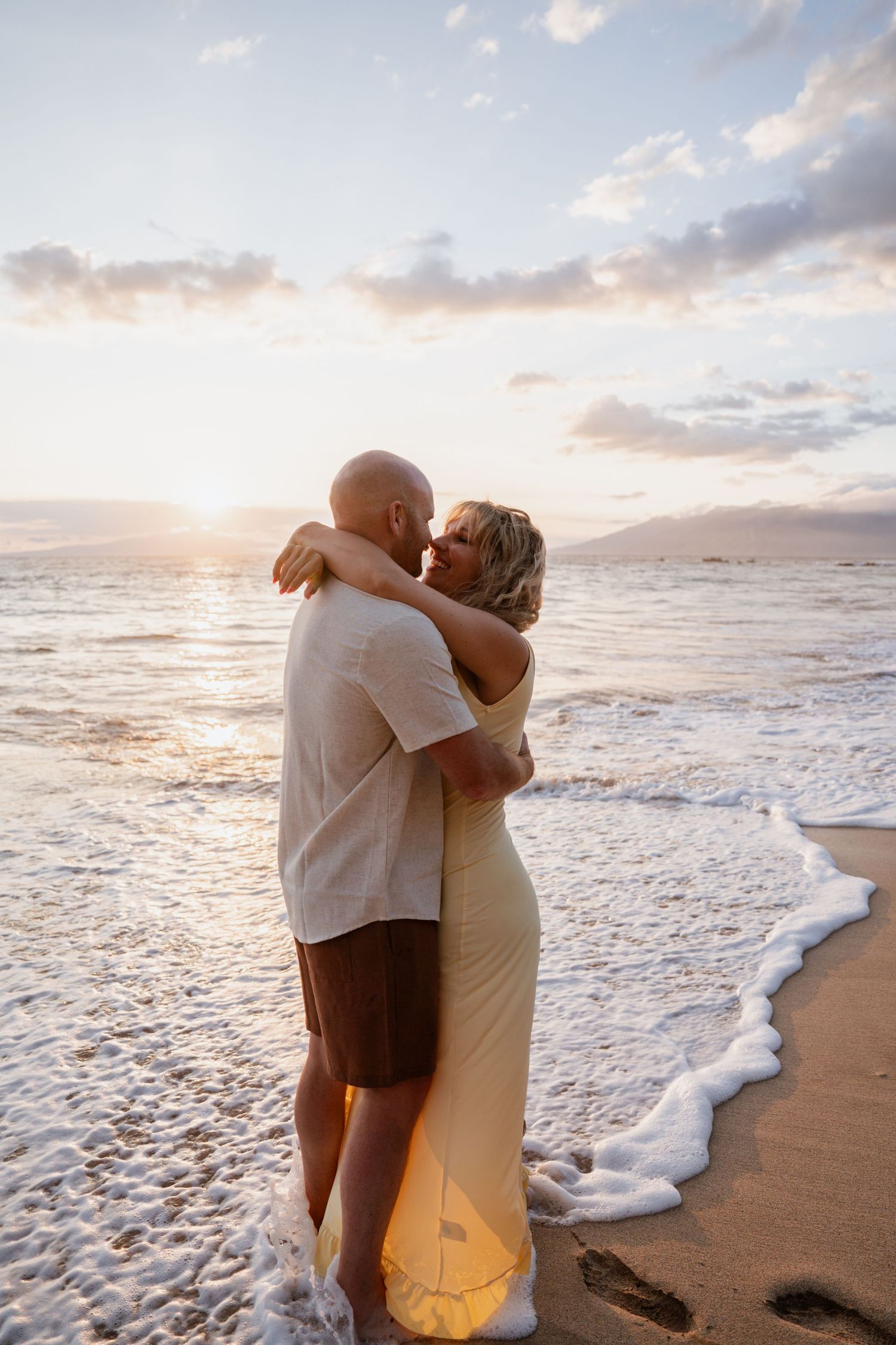 Couple embracing on a sandy beach at golden sunset, waves washing ashore, woman in yellow dress, man in white shirt.