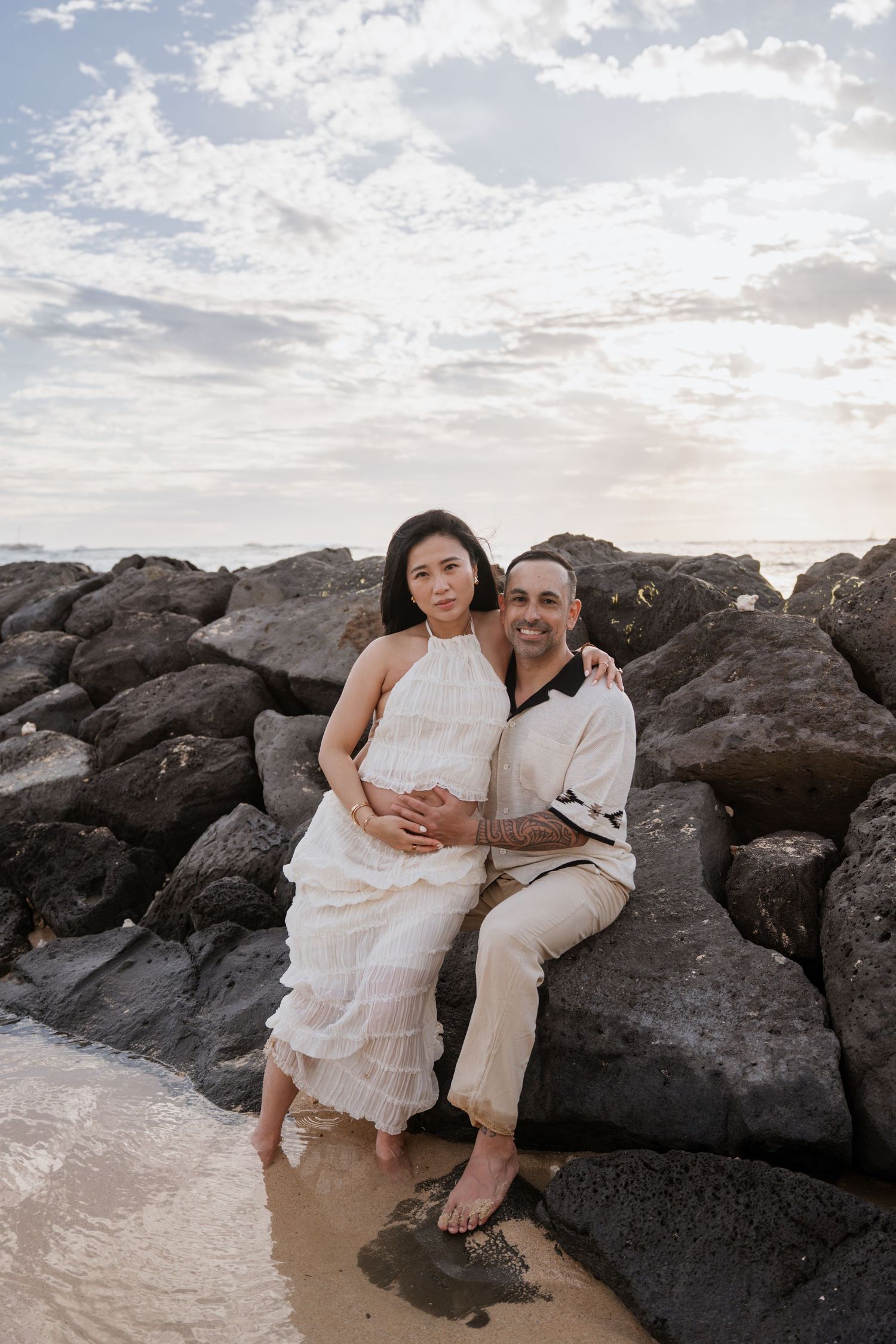 Couple sitting on rocky beach shore at sunset, woman in white dress, man in cream outfit, embracing romantically.