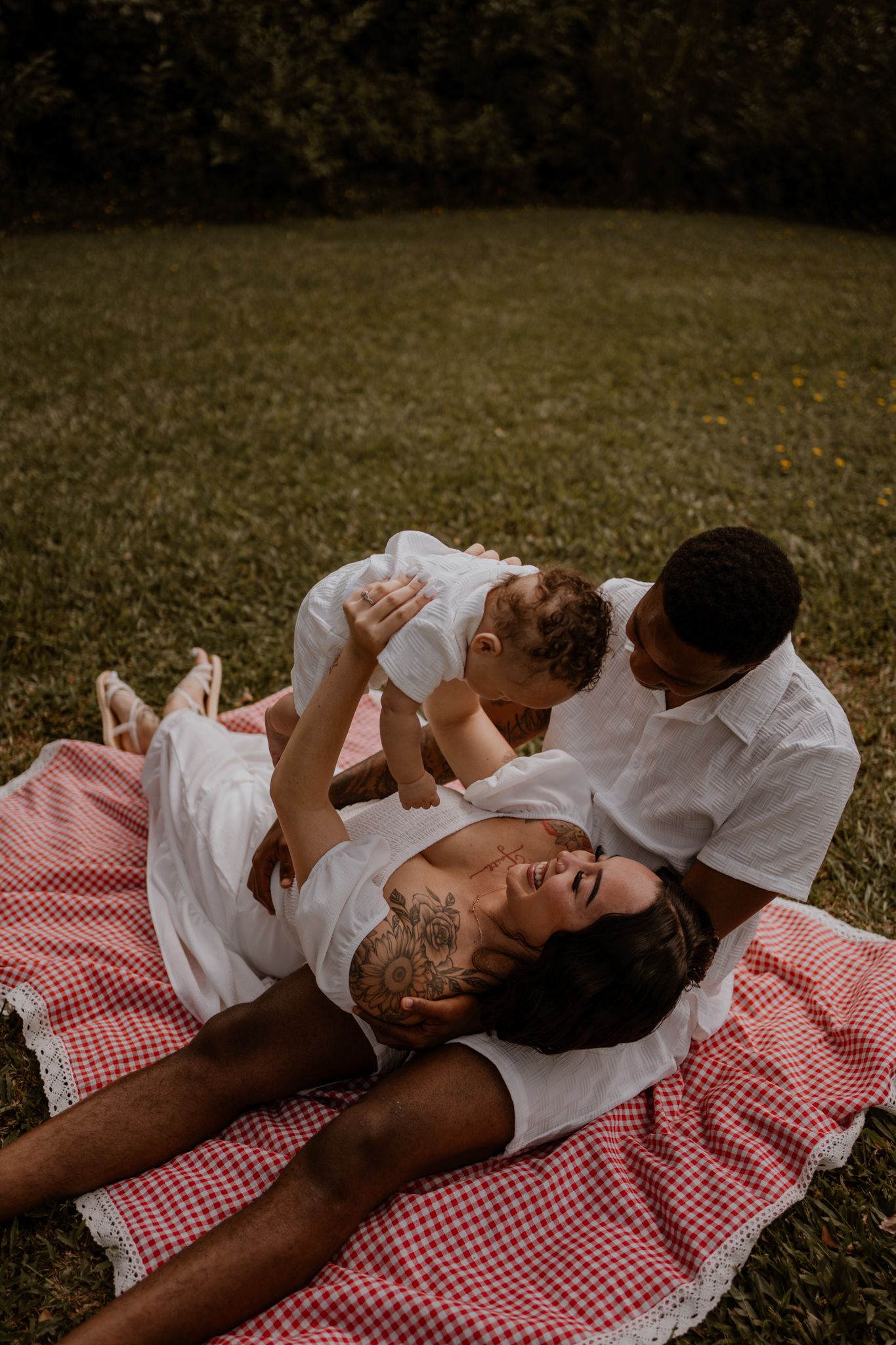 Family shares tender moments while relaxing on a red checkered picnic blanket in a grassy field.
