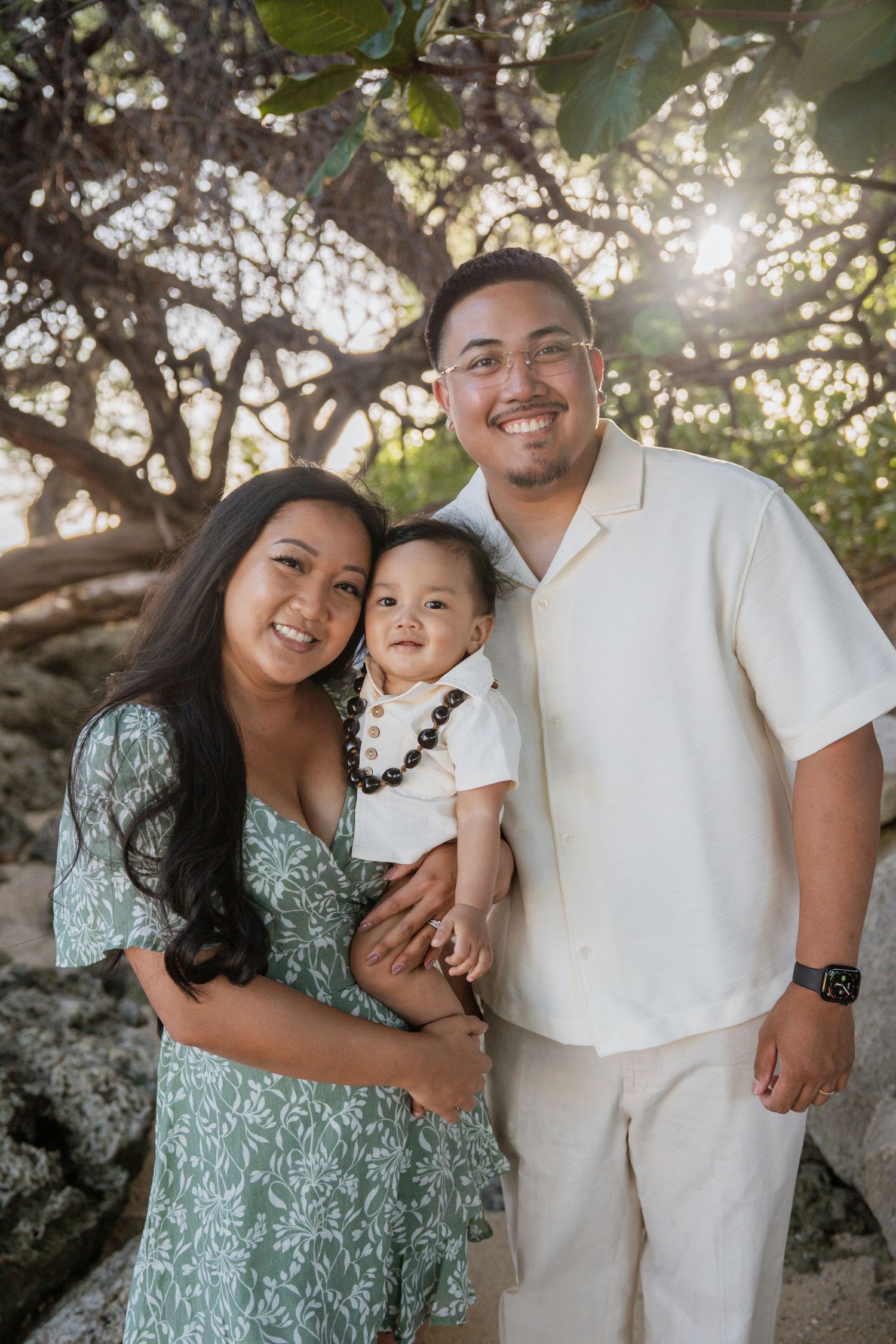 Young family with baby poses outdoors under trees, mom in floral dress and dad in cream shirt, smiling warmly.
