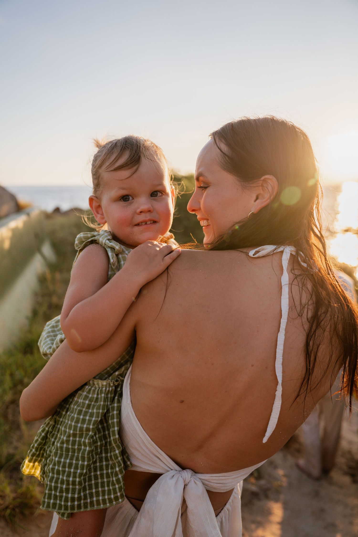 A mother holds her smiling toddler girl at sunset near the ocean, wearing a backless white dress and green checkered outfit.