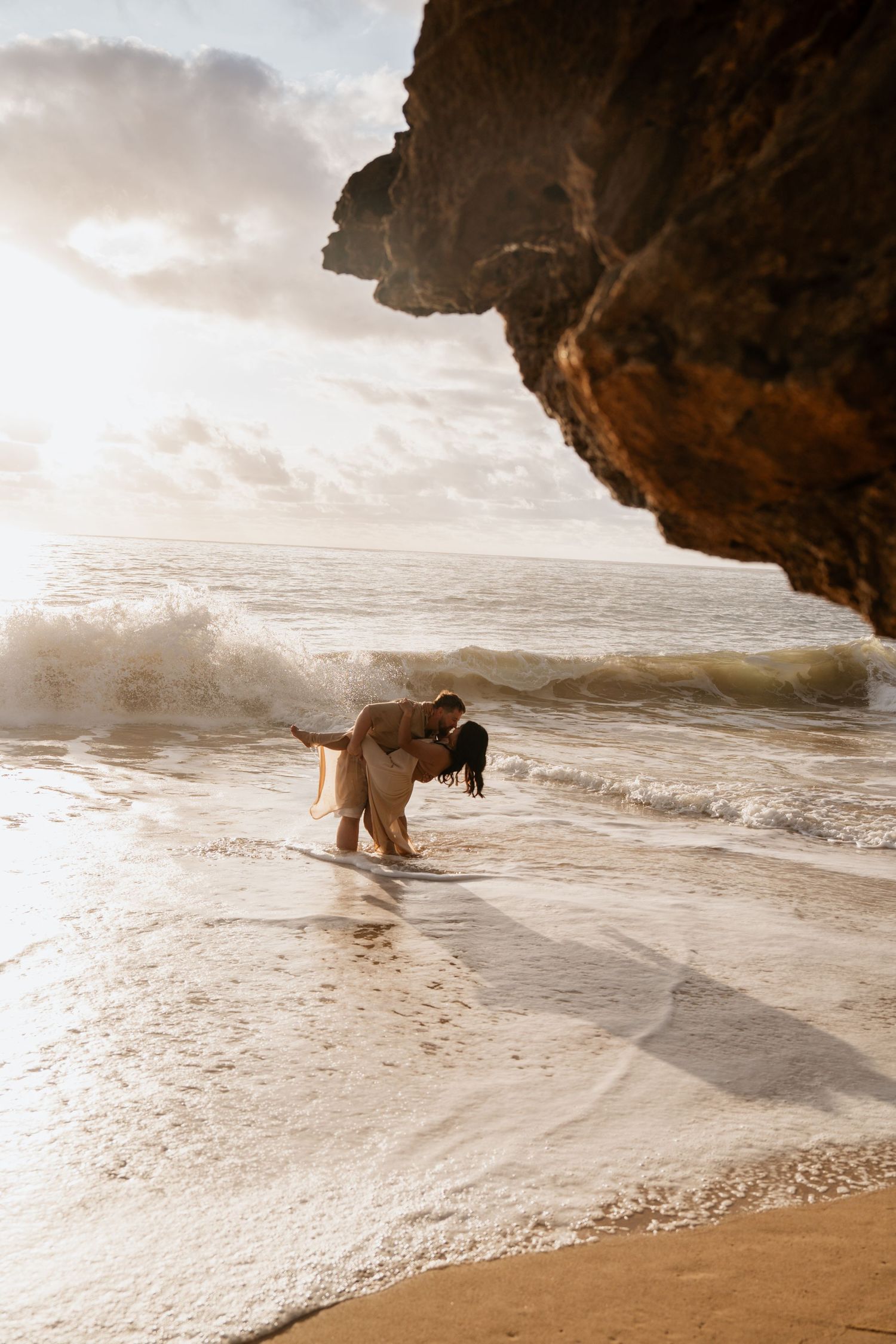 Couple sharing a romantic dip kiss on a sandy beach with crashing waves and dramatic rocky cliffs at golden hour.