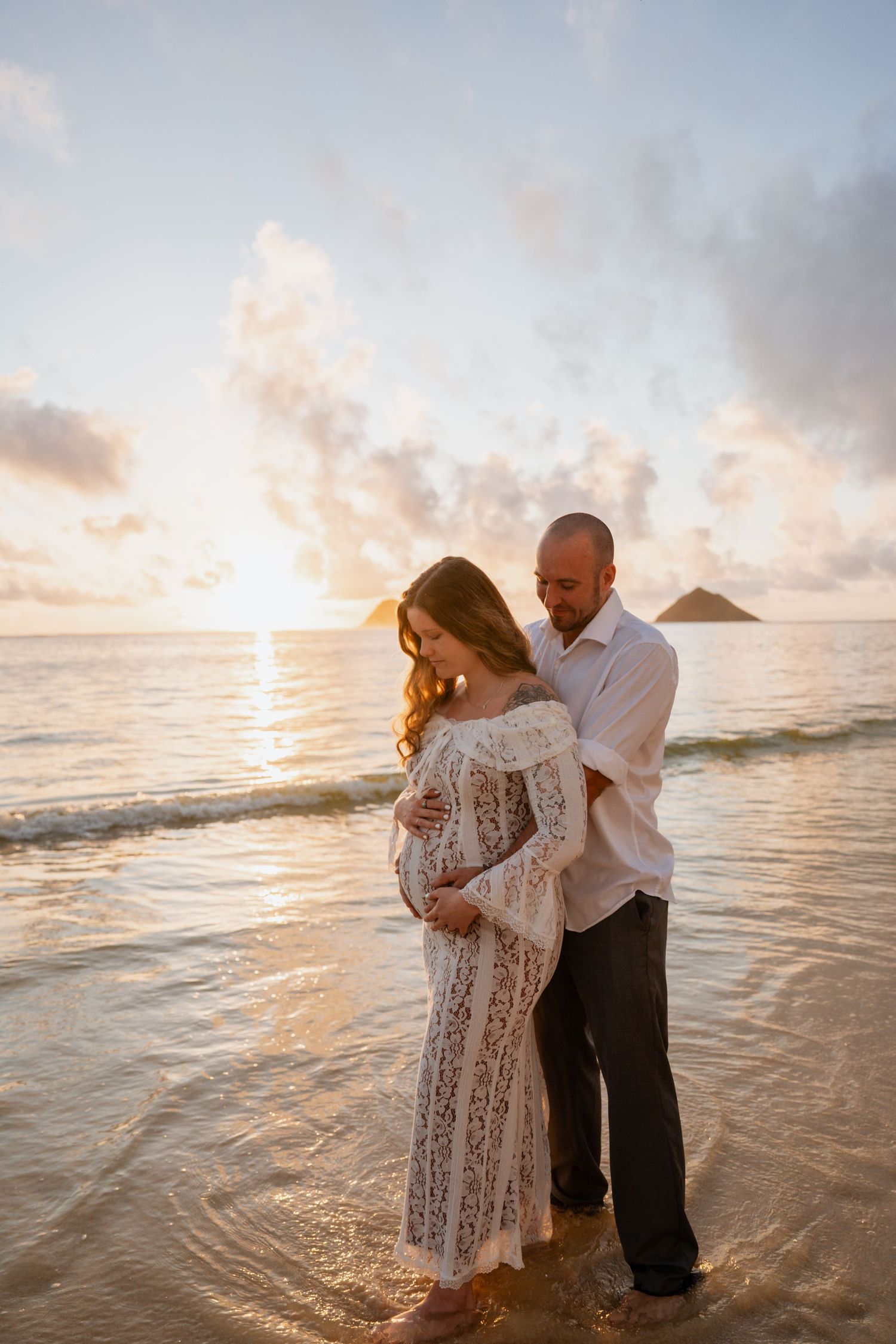 Pregnant couple embracing at sunset on a Hawaiian beach, woman in white lace dress, man in white shirt, ocean waves.