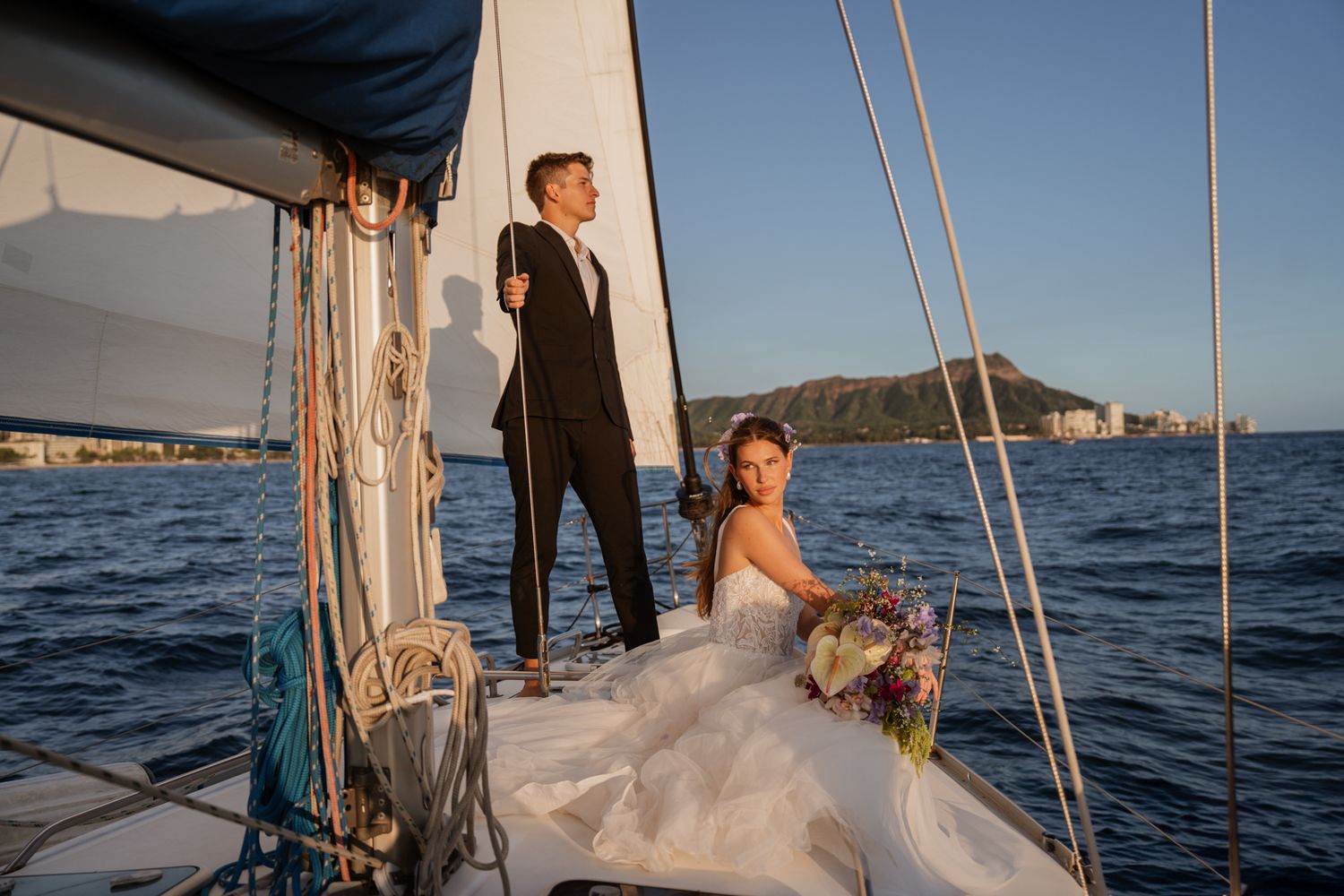 A couple celebrates their wedding day aboard a sailboat on blue waters during sunset near a coastal cityscape.