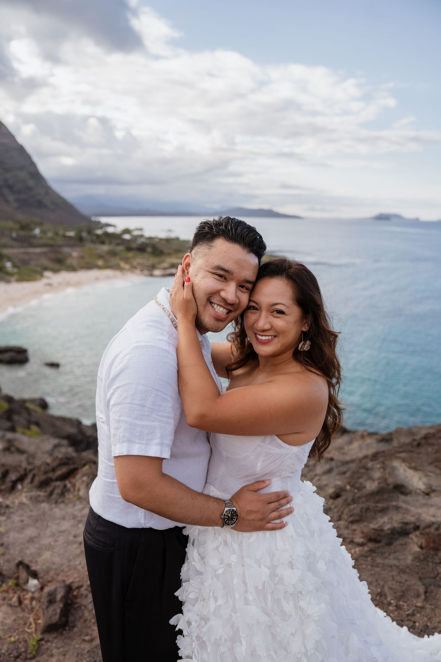 A couple embraces on a scenic rocky coastline overlooking turquoise waters and mountains in Hawaii. Oahu Elopement photographer