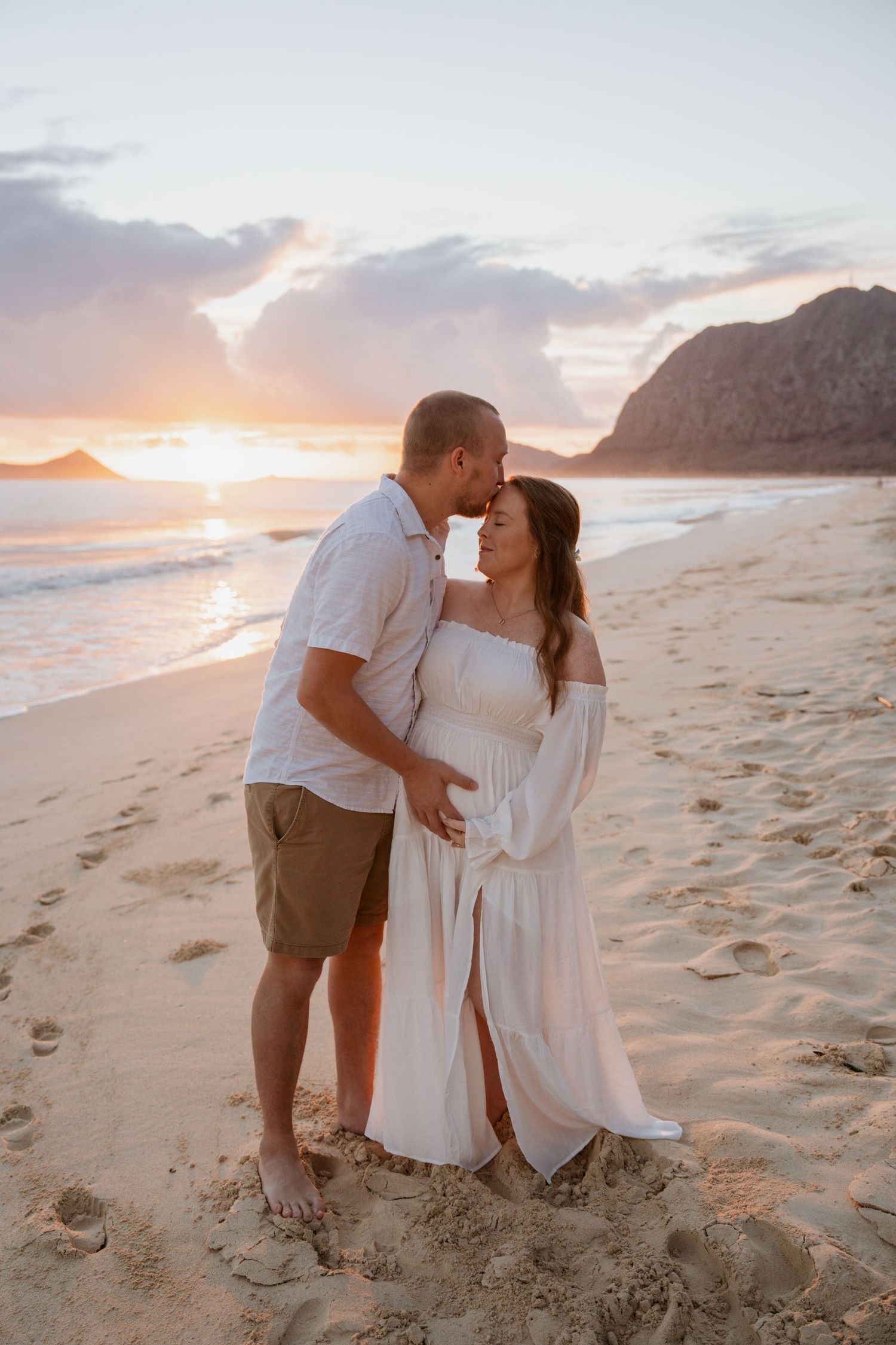 Romantic couple in white outfits sharing intimate moments on a sandy beach at sunset with mountains in background.