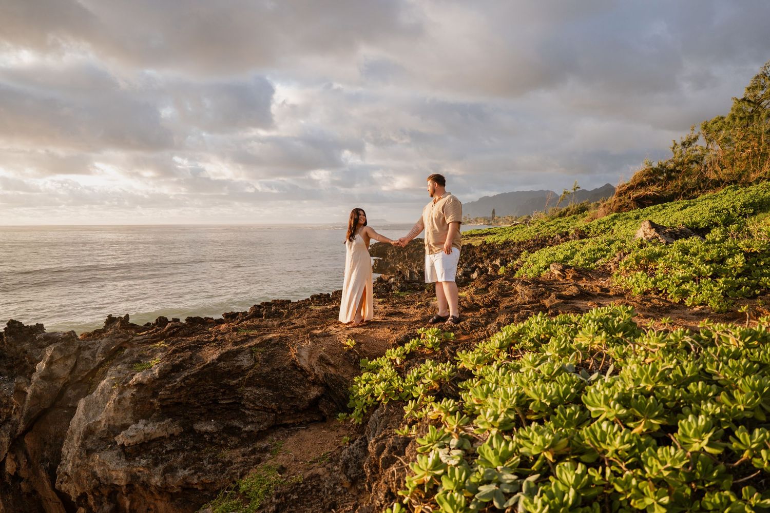 Couple holding hands on rocky Hawaiian coastal cliffside with lush greenery and ocean views at golden sunset.