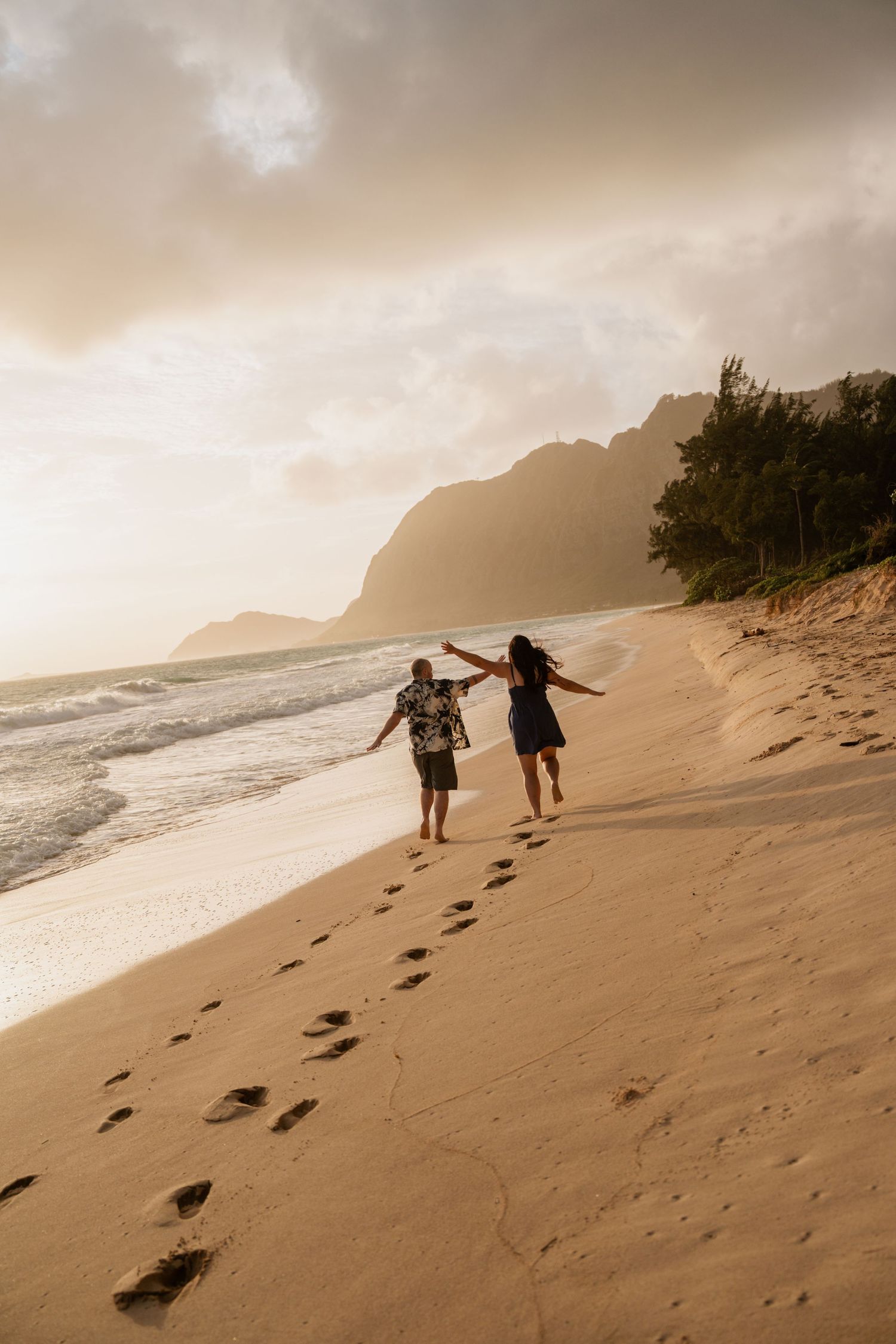 Couple holding hands walking along a sandy beach at sunset with dramatic mountain cliffs and golden sky.
