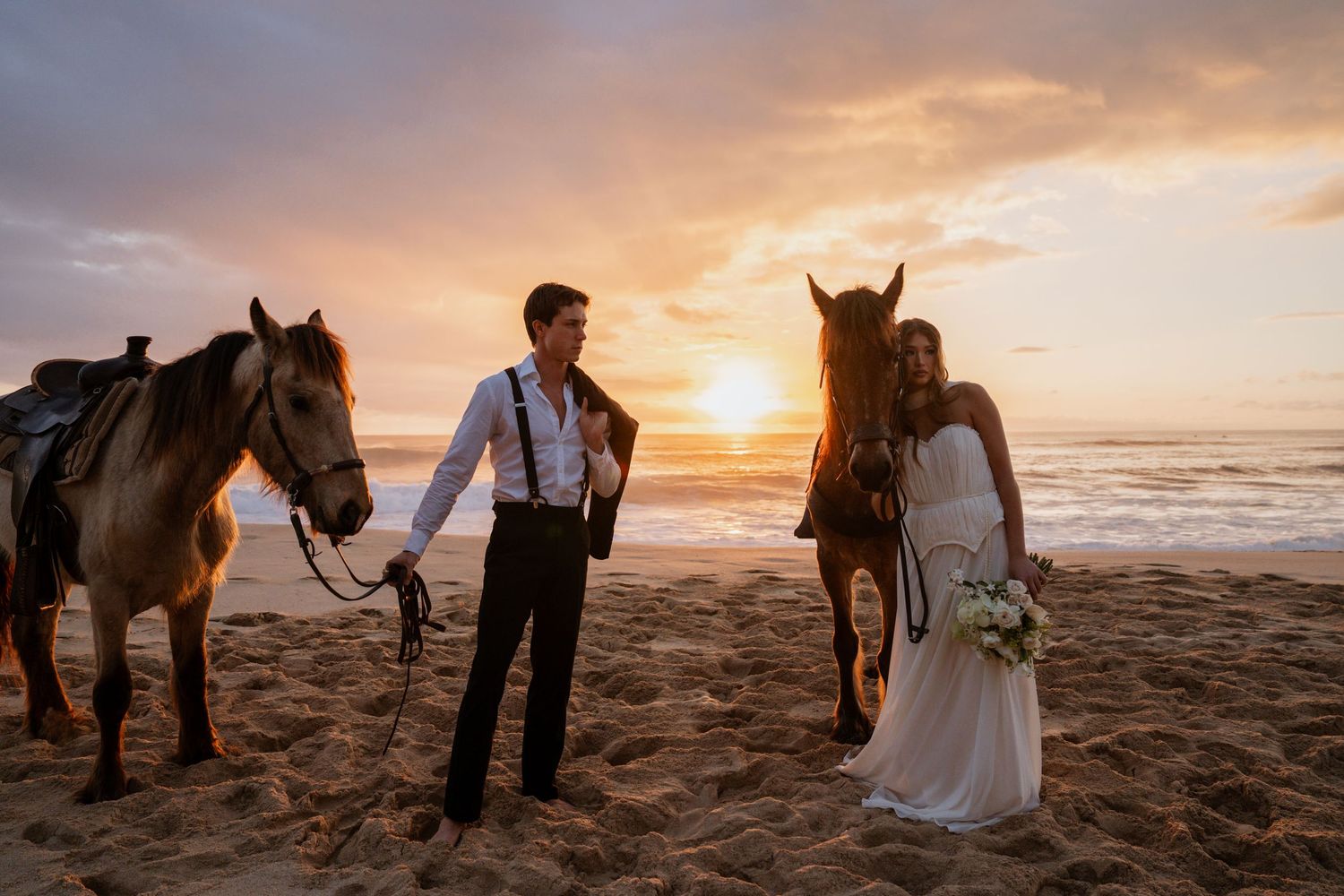 A romantic beach wedding photoshoot with horses during a golden sunset on the sandy shore.