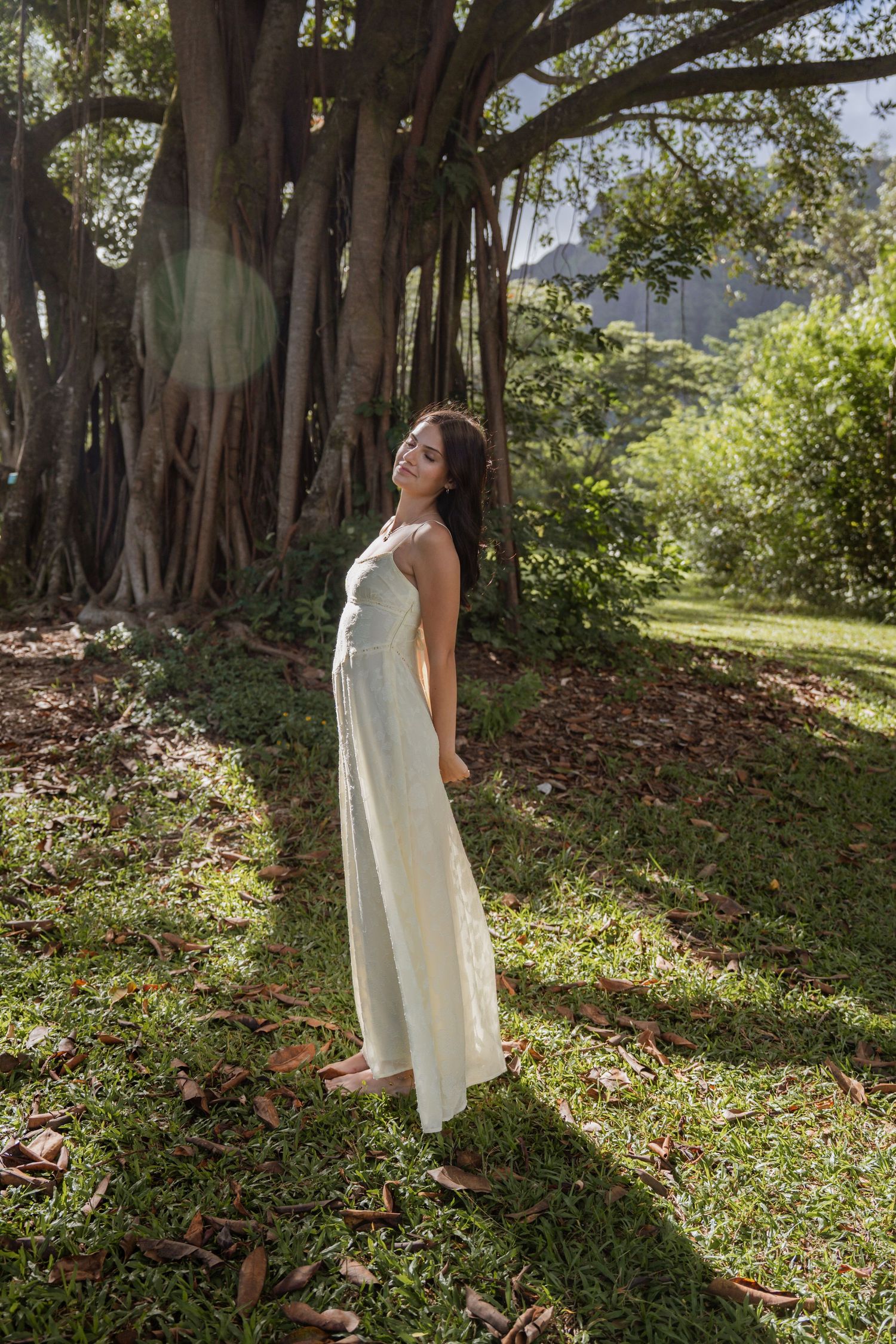 A person in a flowing white maxi dress poses in a sunlit garden with large trees creating a natural backdrop. Senior graduation photoshoot