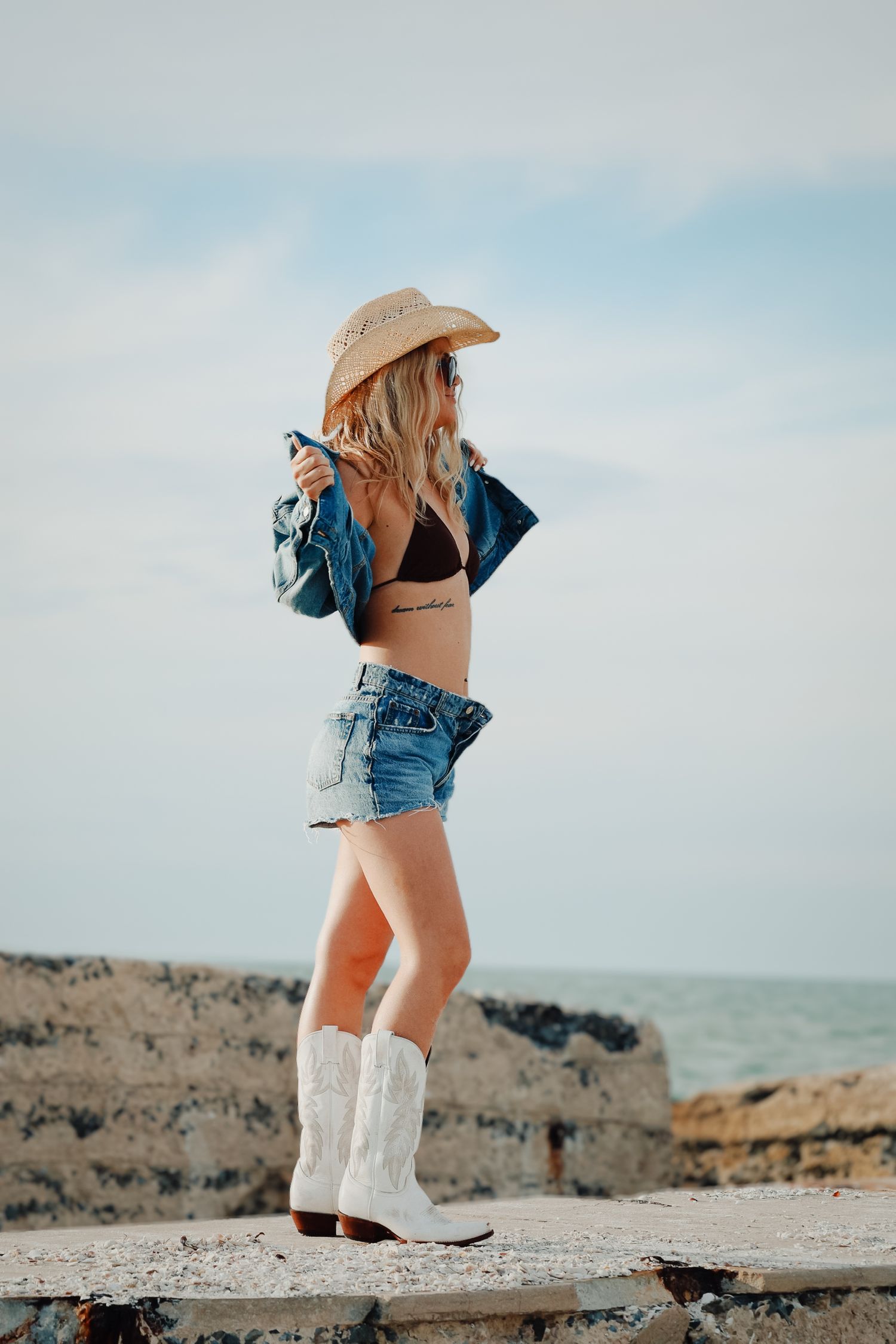 Woman in denim outfit and white boots poses on rocky beach overlook.