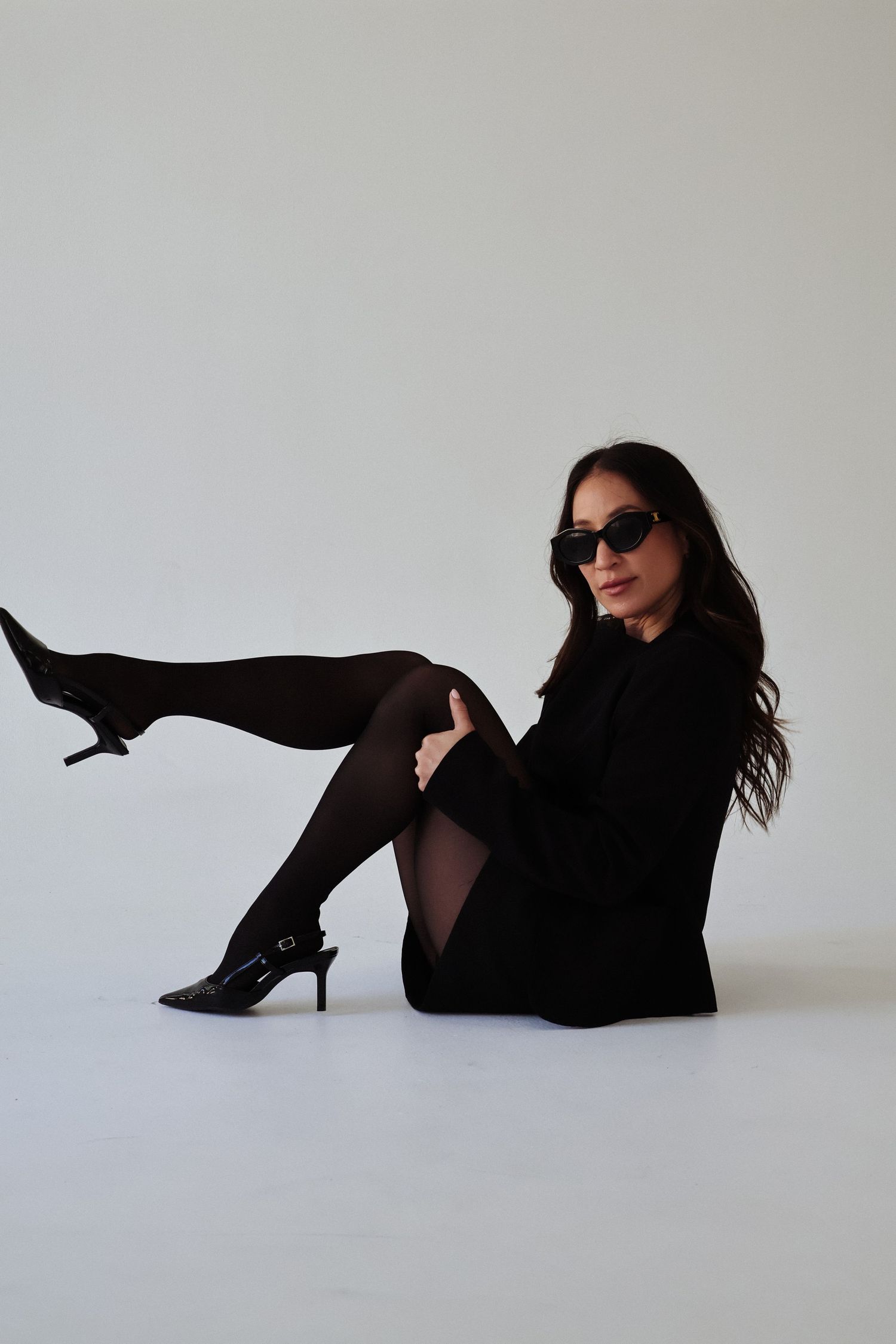 Fashion model in black outfit and sunglasses poses dynamically on white background.