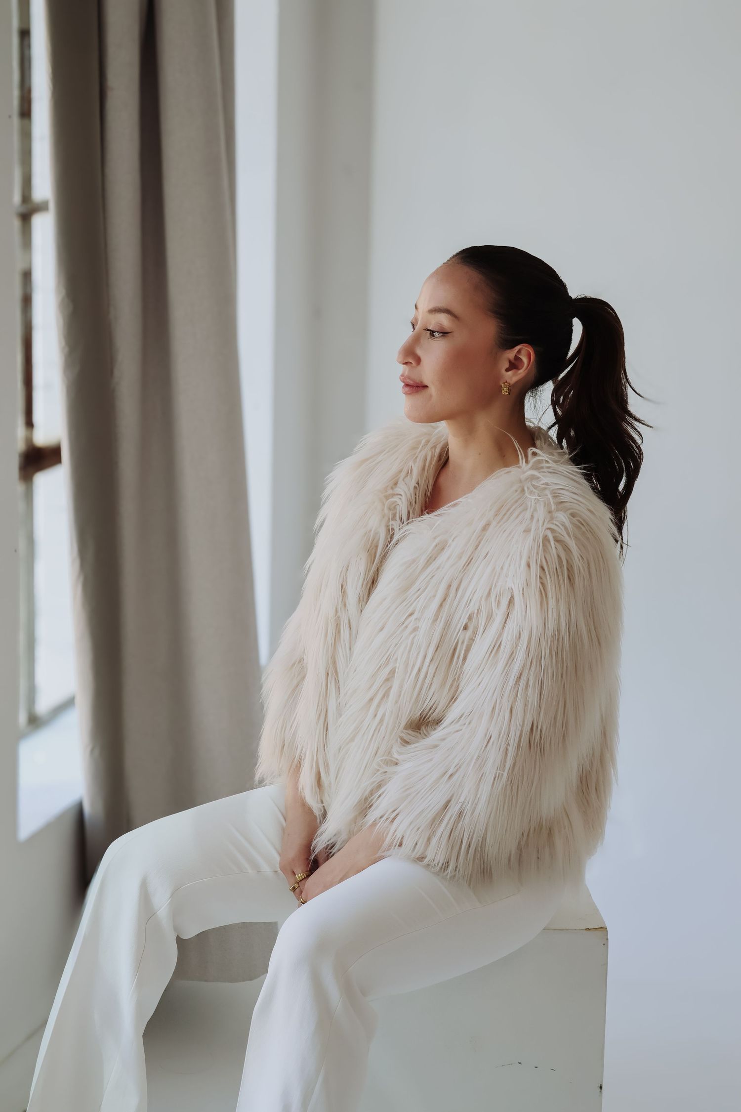 Model in cream faux fur coat and white pants poses by window.