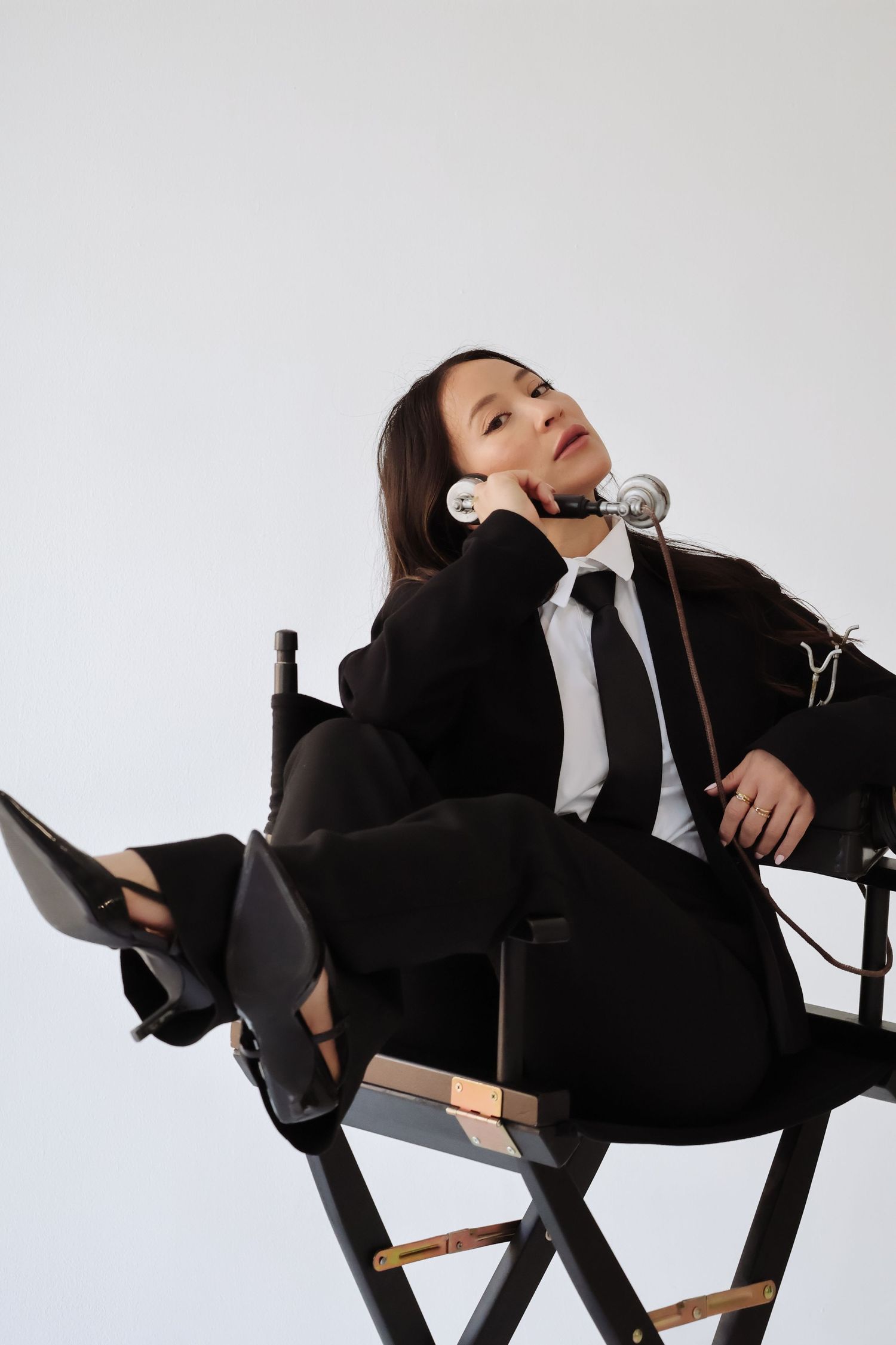 Professional model in black suit and tie reclines on director's chair during studio photoshoot.