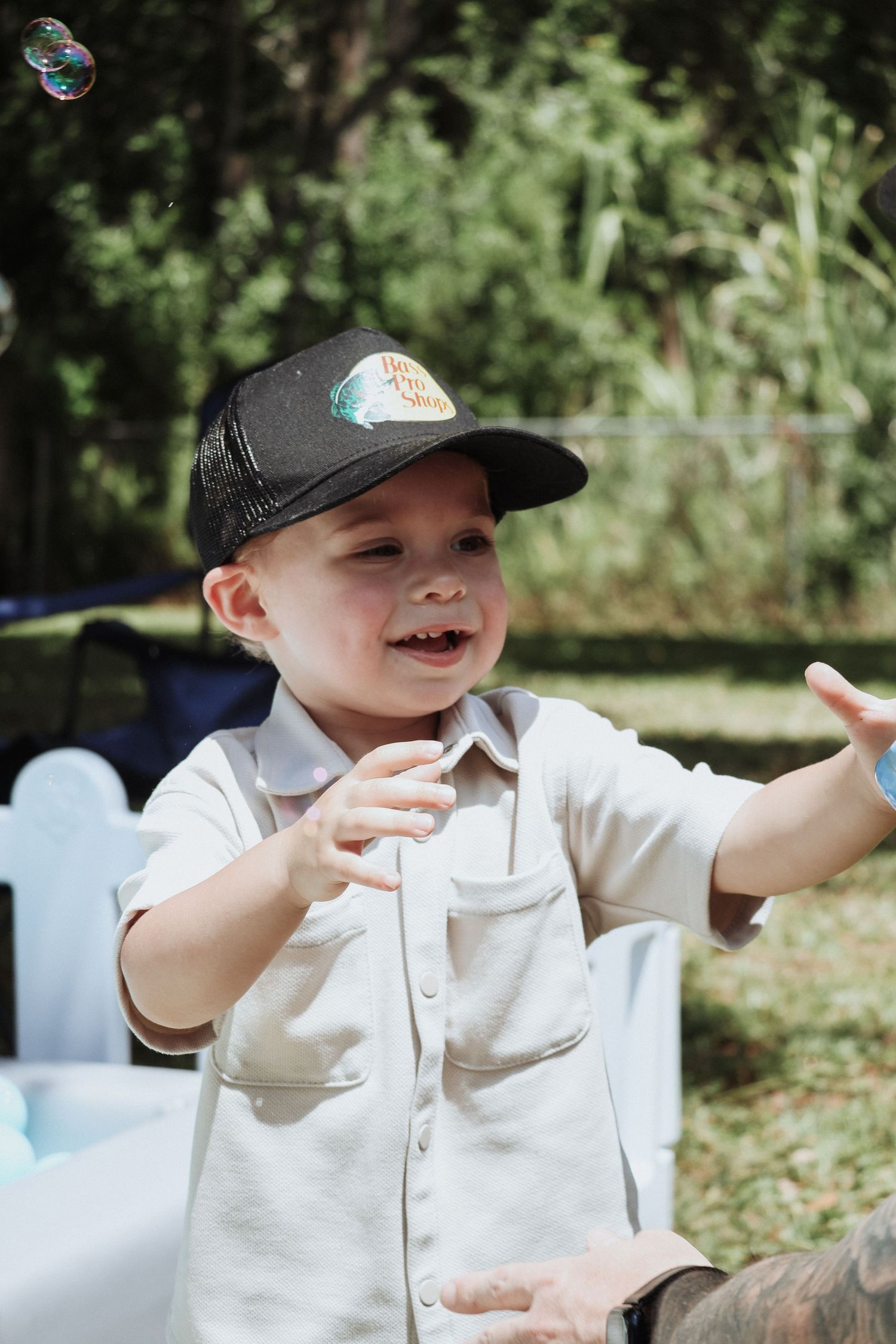 Child in white shirt and baseball cap reaching for bubbles outdoors.