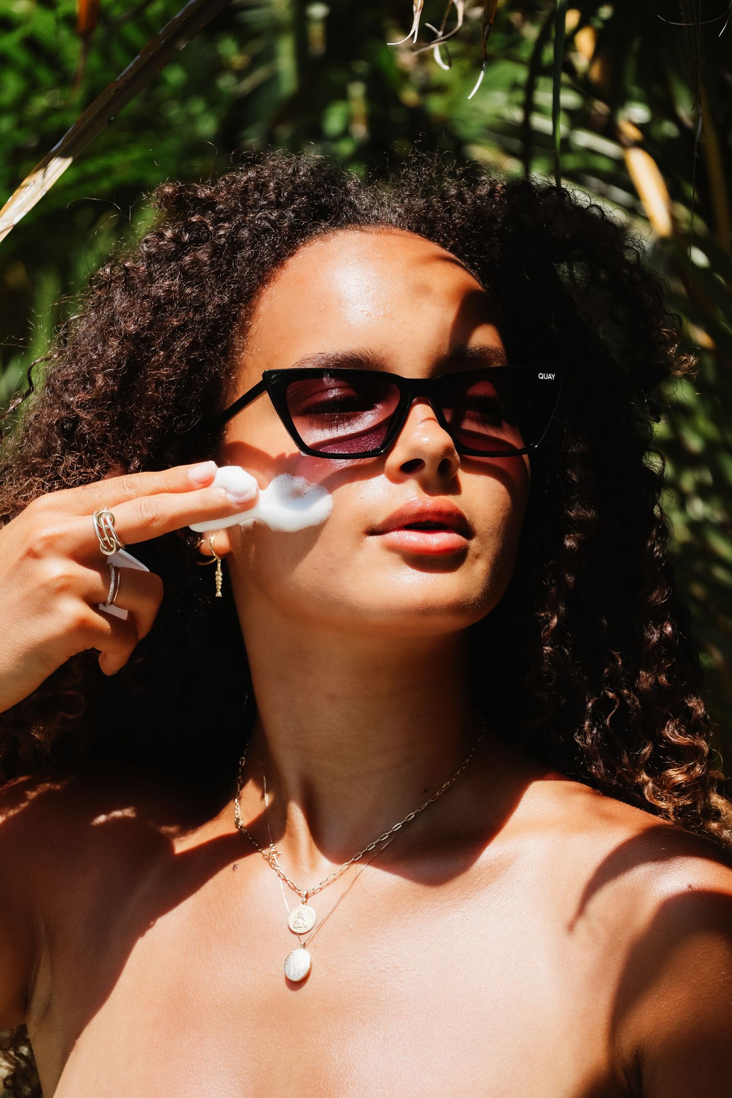 Fashionable natural hair model wearing sunglasses and necklace poses in sunlit garden setting.