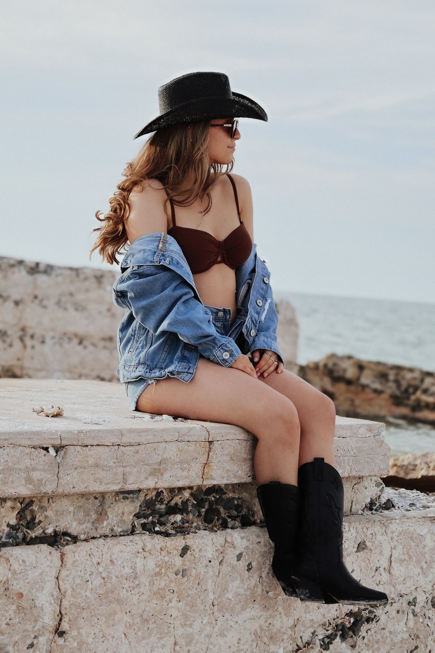 Beachwear fashion with brown bikini, denim jacket and black cowboy hat against coastal scenery.