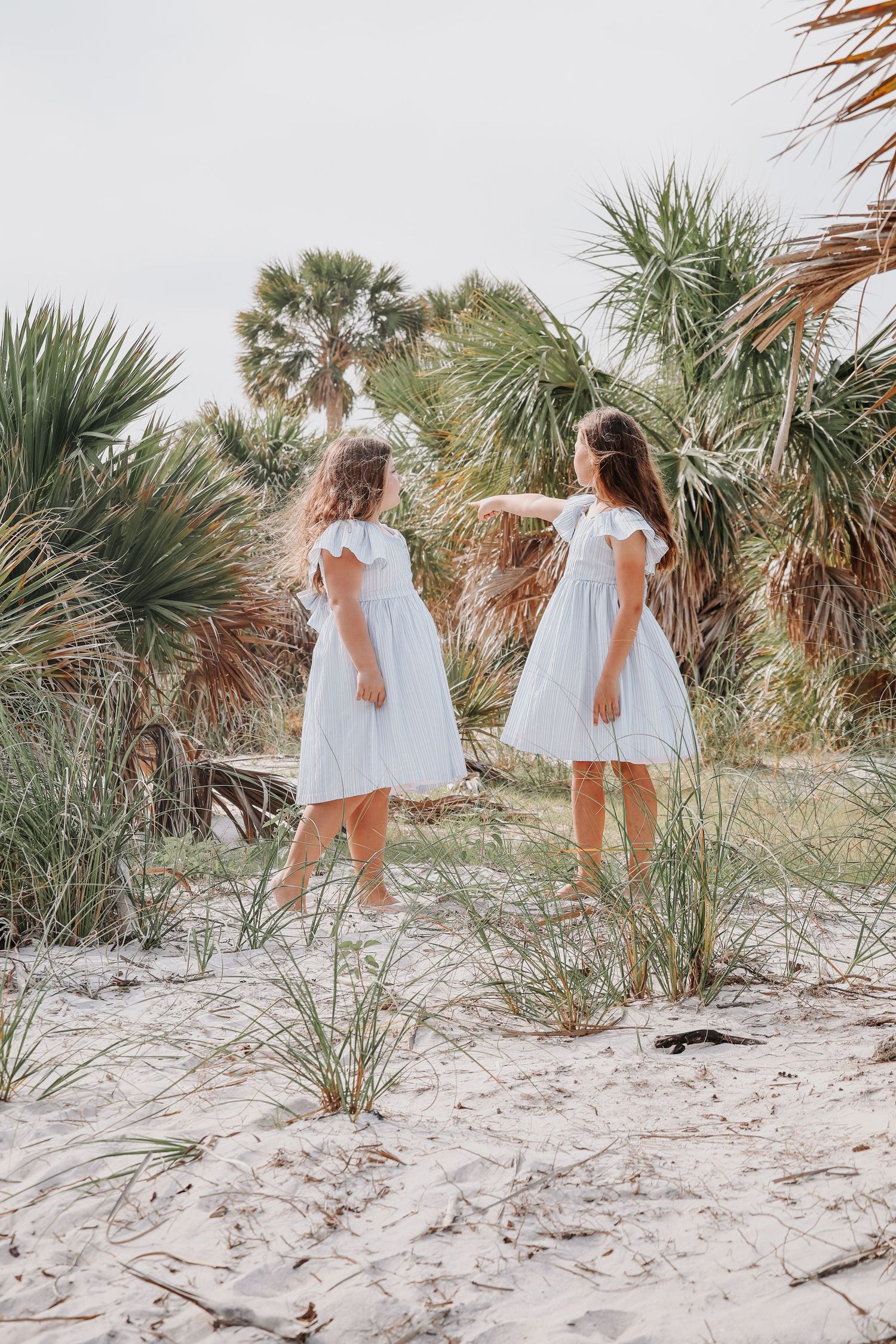 Two people in white summer dresses pose together on sandy dunes with tropical plants in background.