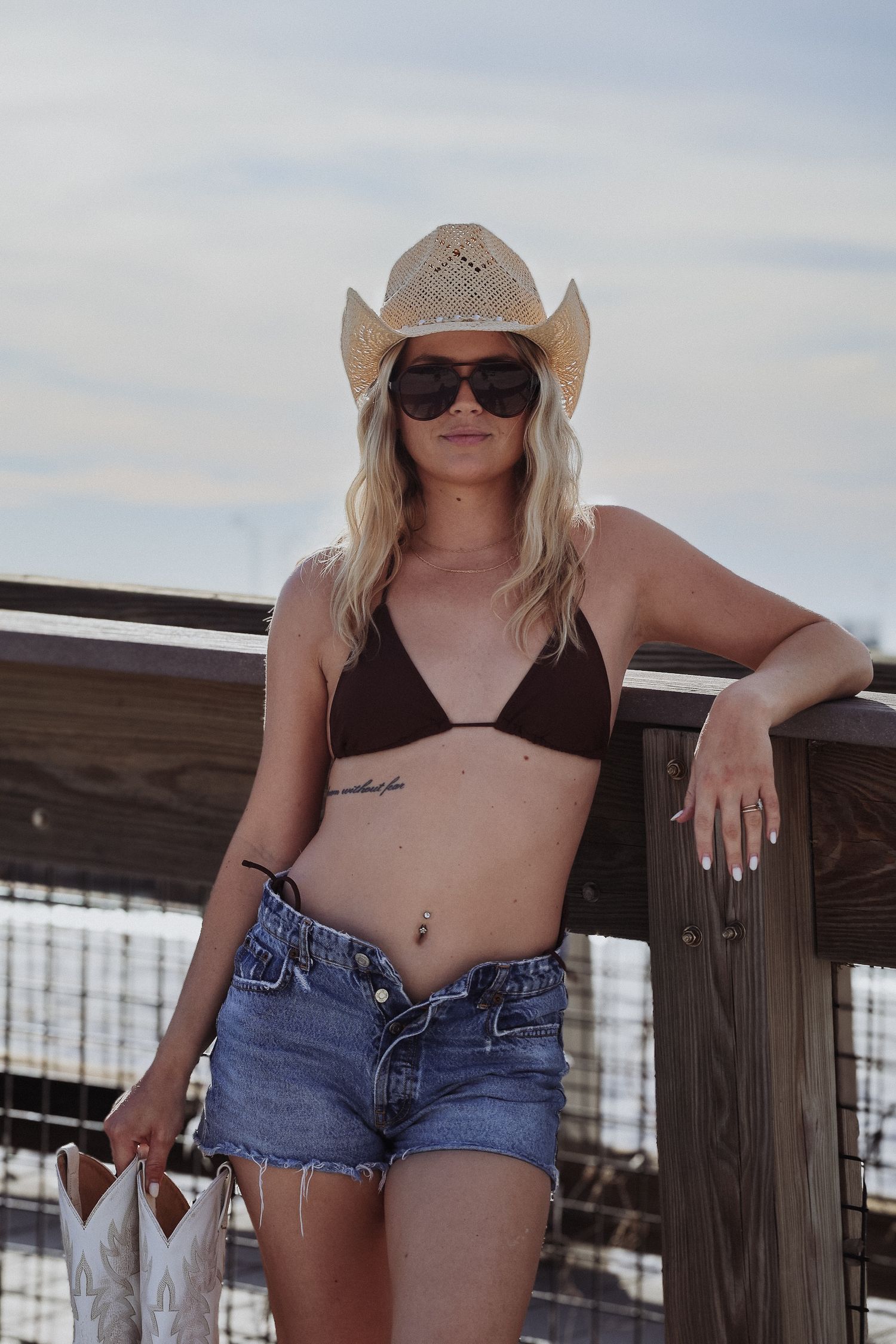 Casual beach look with straw cowboy hat, bikini top and denim shorts against wooden railing.