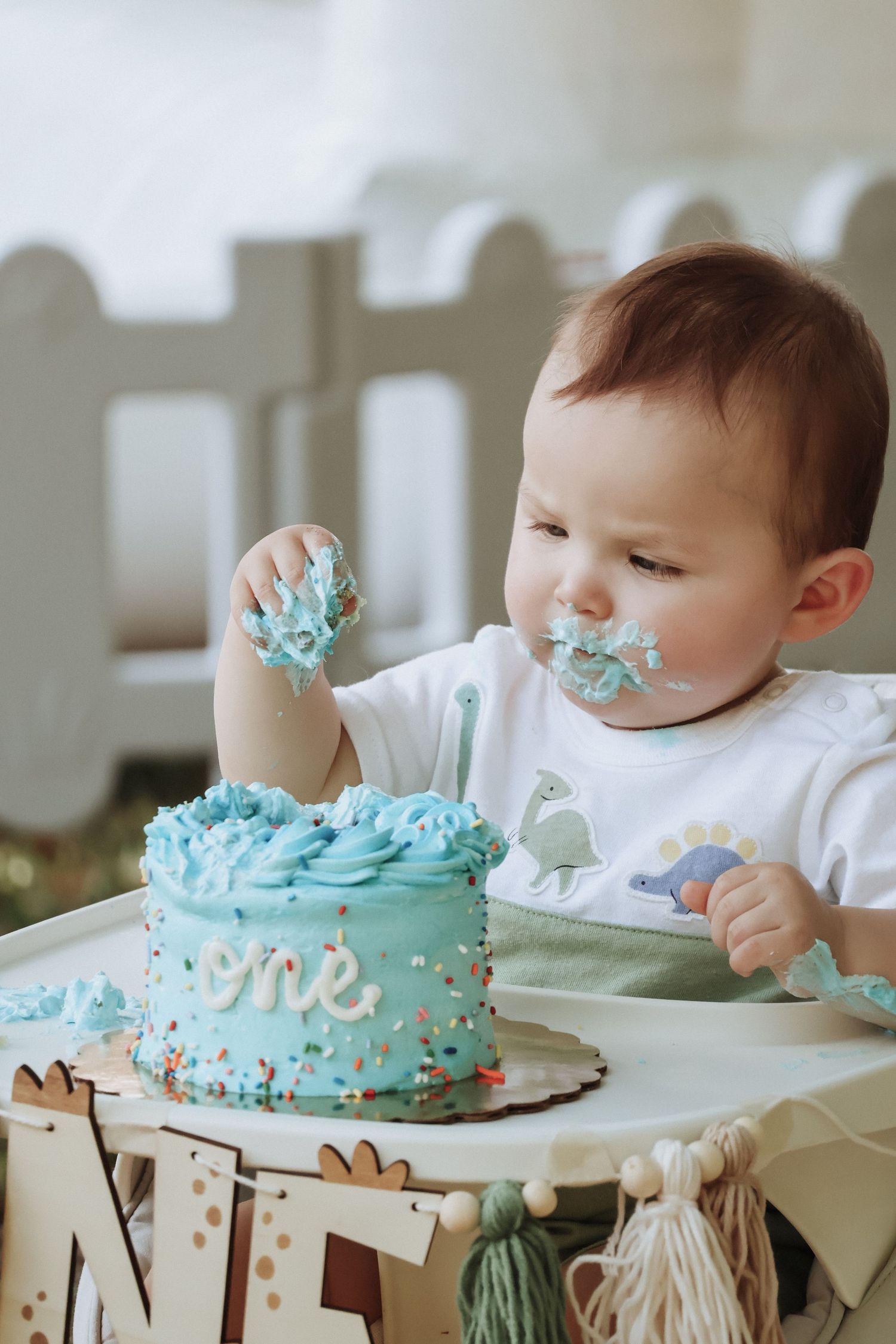 Toddler with frosting-covered face reaches for blue birthday cake decorated with sprinkles.