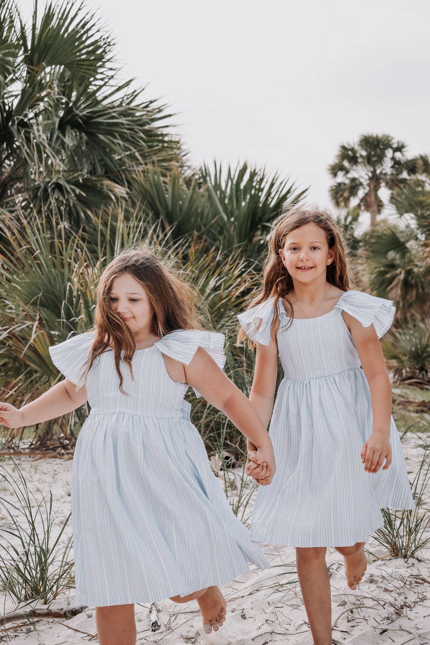 Two people in white dresses hold hands while walking through palm trees and sand dunes.