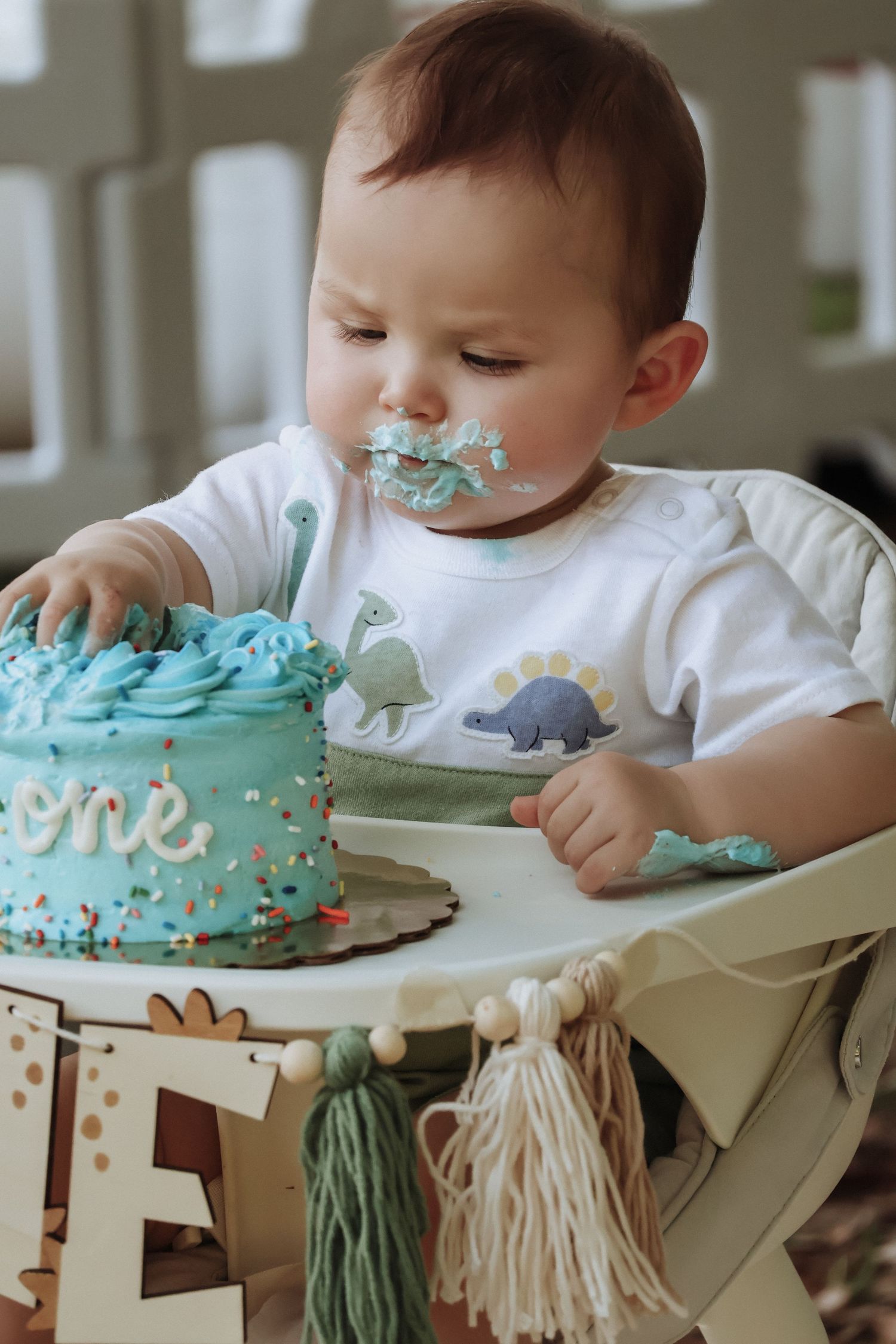 Baby wearing dinosaur shirt enjoys a blue frosted smash cake during first birthday celebration.