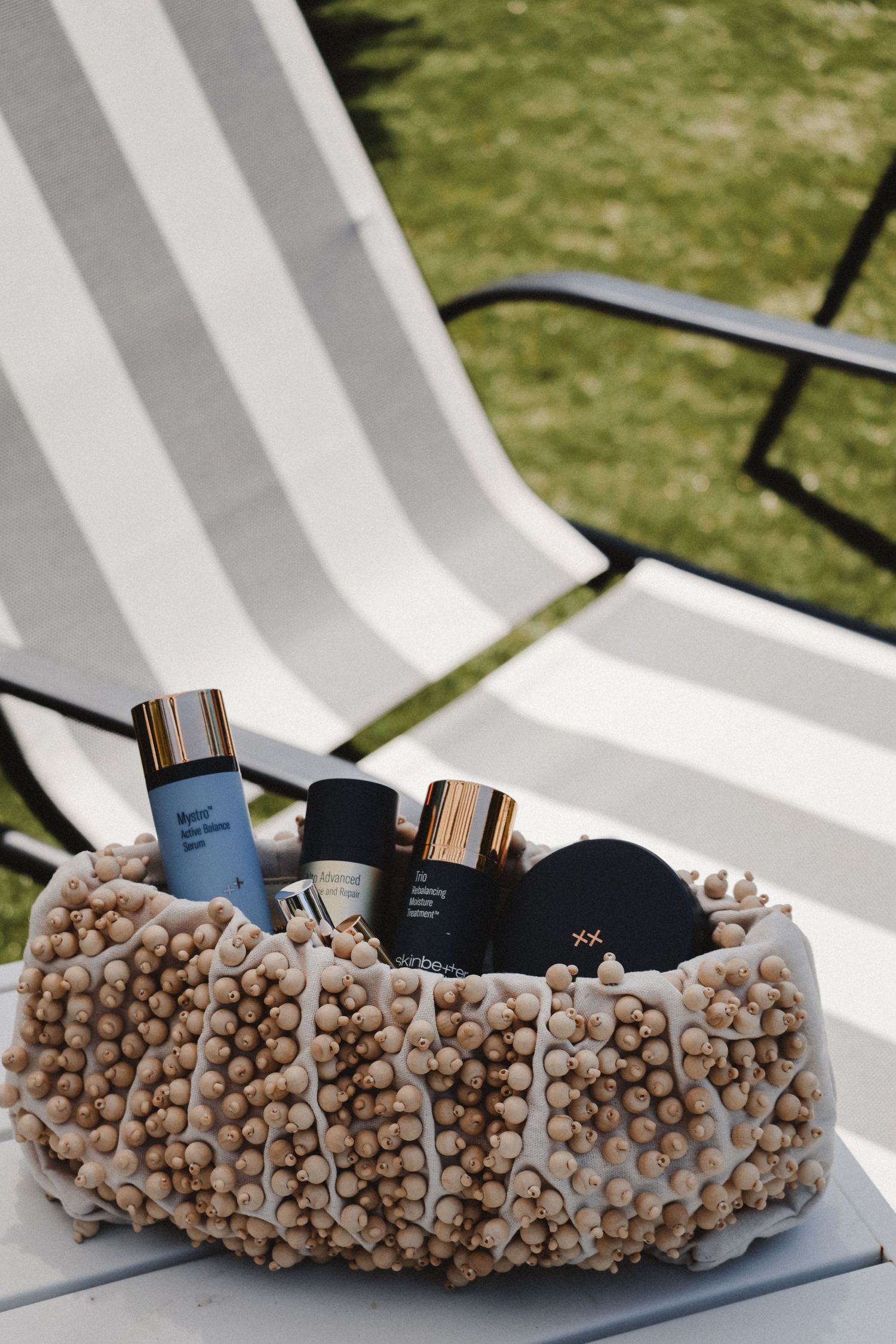 Beaded beach bag with blue cosmetic bottles displayed on white lounge chair outdoors.
