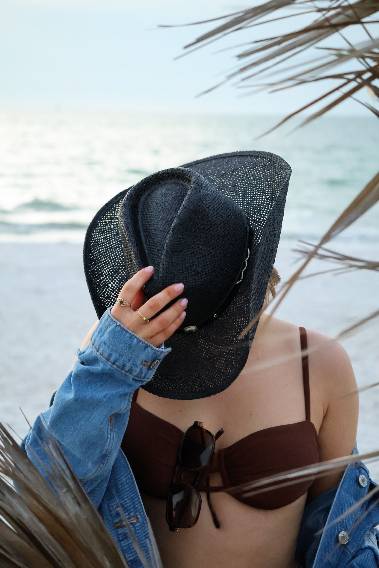 Artistic beach photo of person in denim jacket holding black cowboy hat.