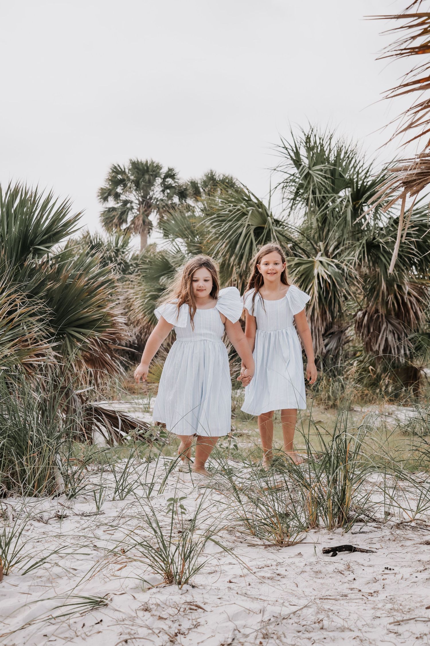 Two people in matching white sundresses stand together on a sandy beach path surrounded by palm trees.